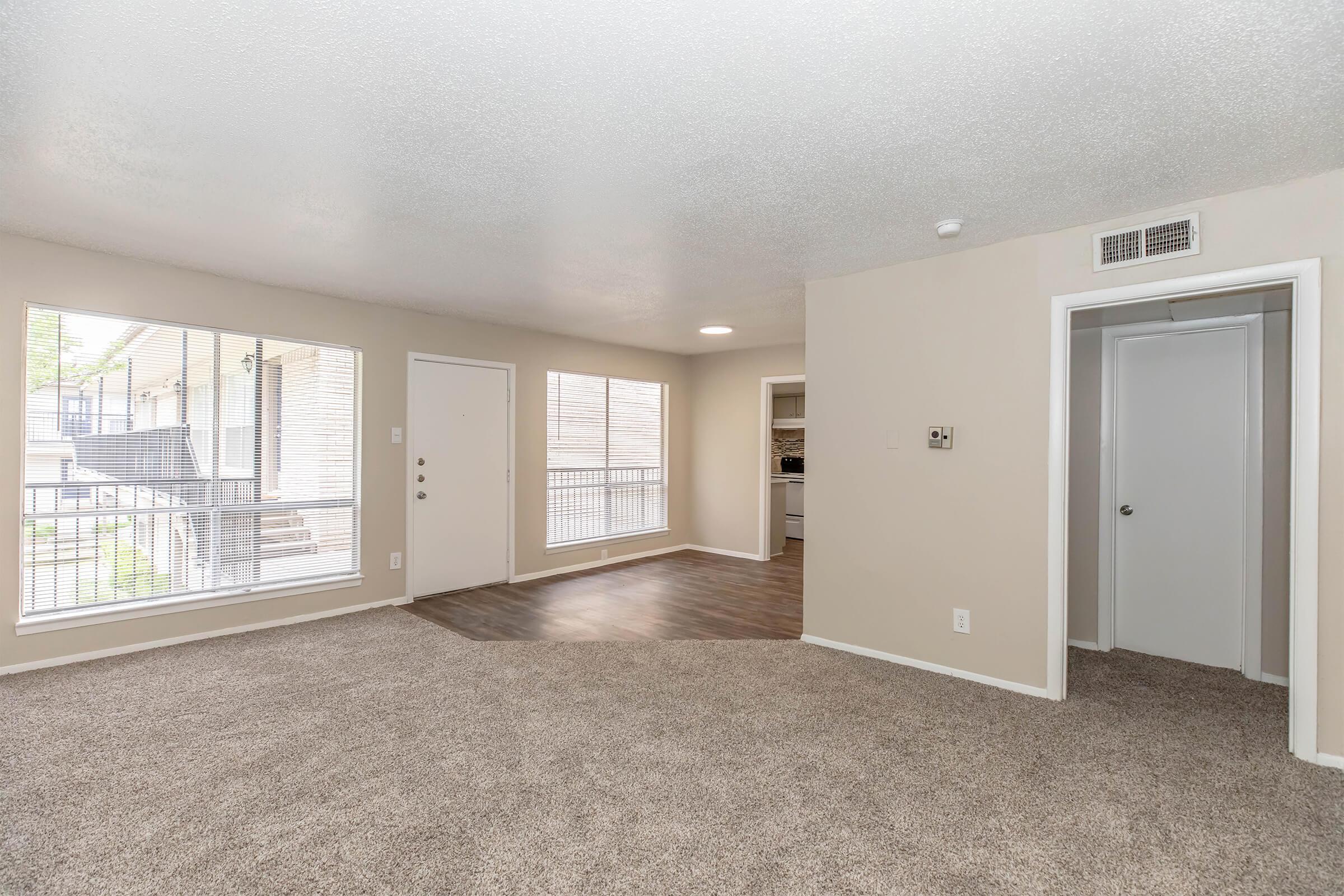 Spacious interior of a well-lit apartment featuring a carpeted living area with large windows. A door leads to a balcony. The room connects to a kitchen visible through an open doorway and includes a closet on the right, enhancing the apartment's functionality and inviting atmosphere.