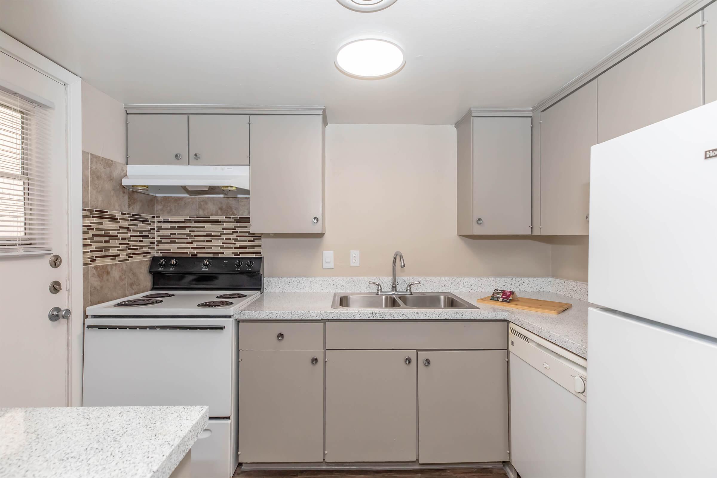 A modern kitchen featuring light gray cabinets, white appliances, a tile backsplash, a double sink, and a granite countertop. The kitchen includes a stove, refrigerator, and dishwasher, with natural light coming through a window and door.