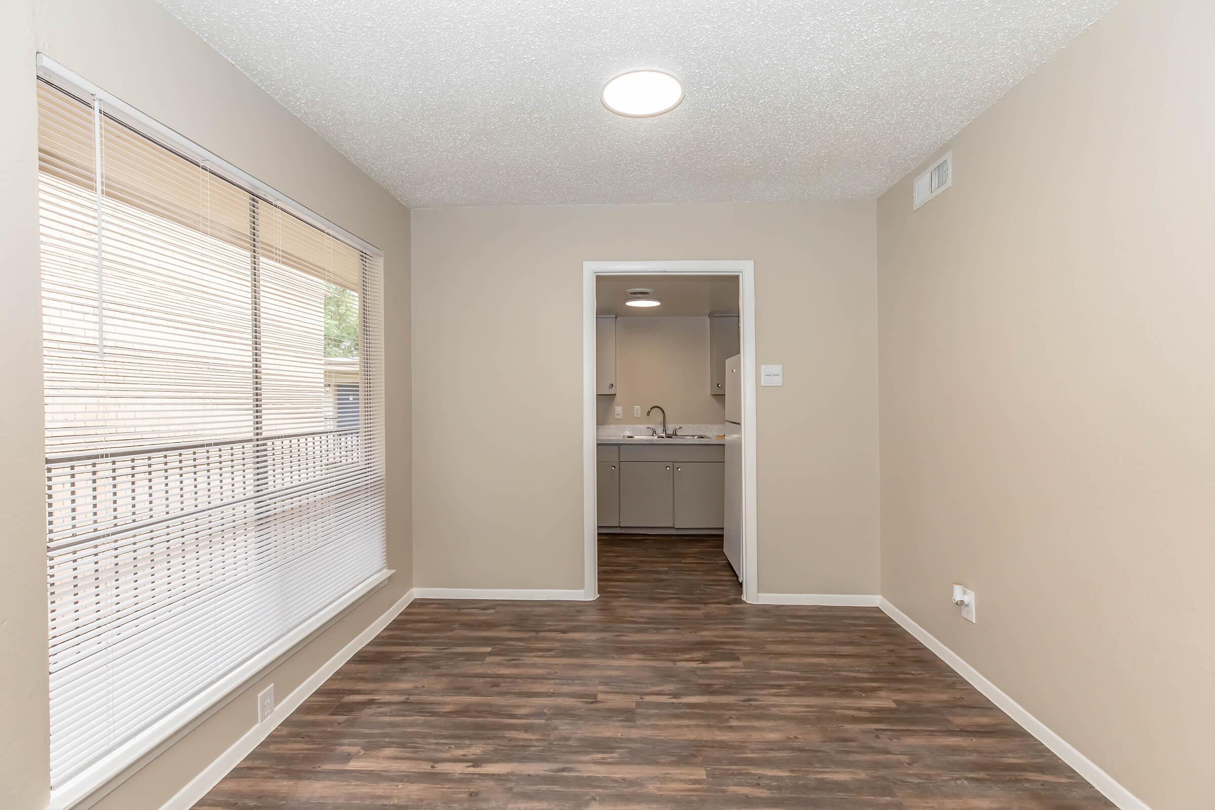 A well-lit, empty room featuring light-colored walls, a textured ceiling, and wooden flooring. A large window with blinds allows natural light in, and a doorway leads to a bathroom with a double sink visible in the background.