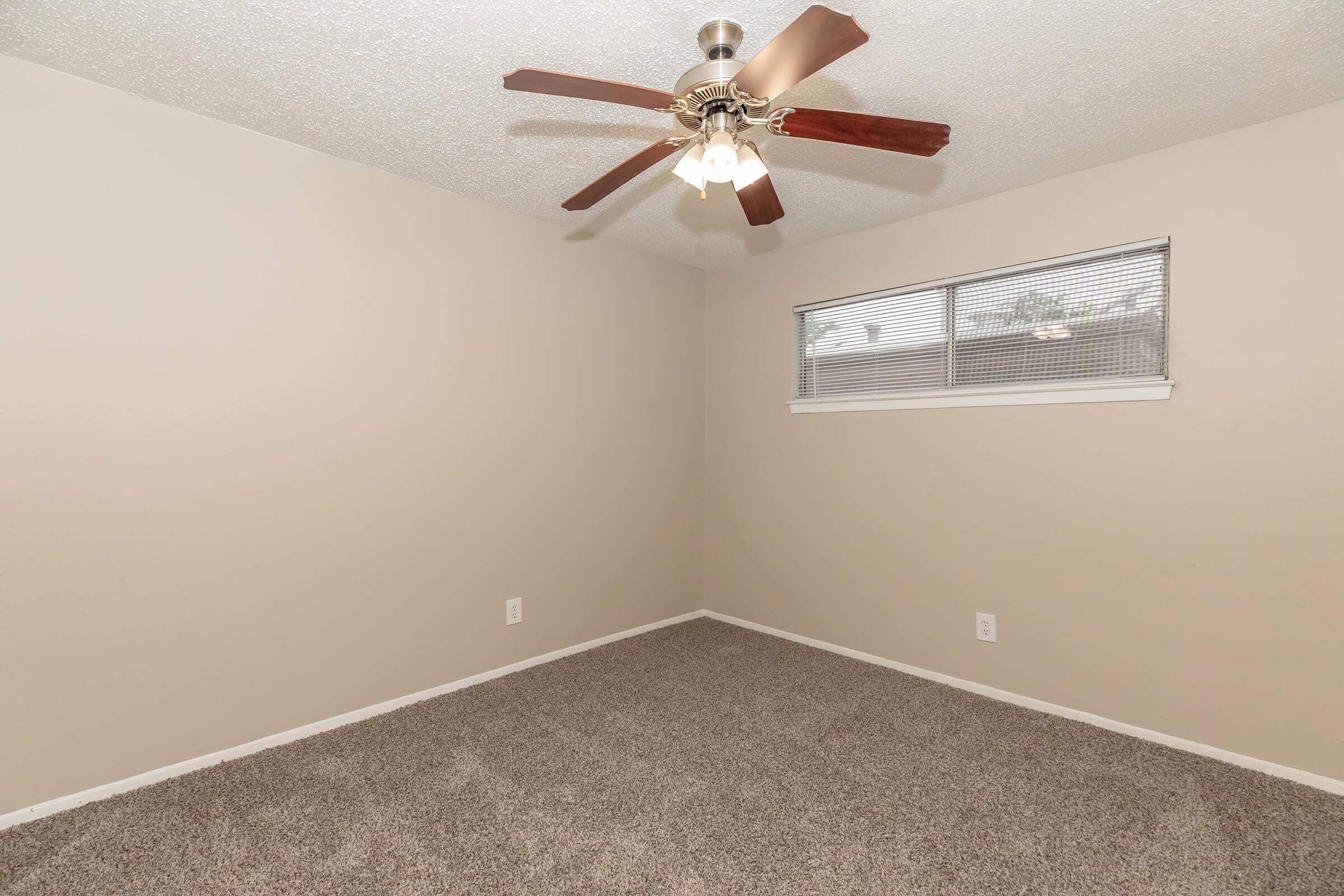 A vacant room with beige walls and carpeted flooring. A ceiling fan with light fixtures is mounted on the ceiling. A window with blinds is visible on one wall, providing natural light. The space appears neat and ready for furnishing.