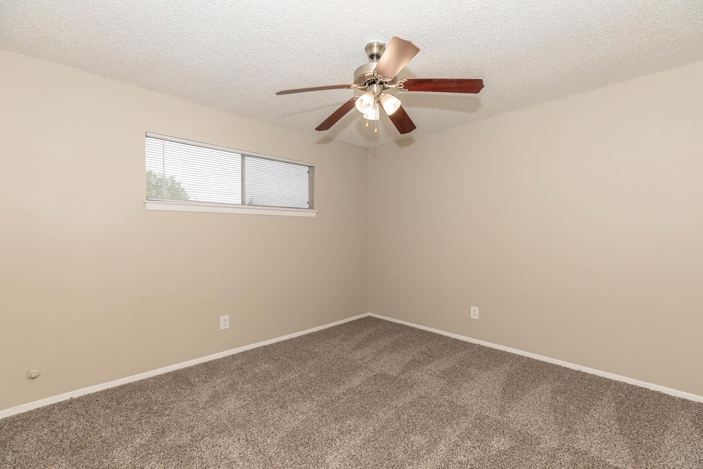 Empty room featuring beige walls and a light brown carpet. The room includes a ceiling fan with four lights and a small window with blinds, allowing natural light to enter. The space is clean and unfurnished, providing a blank canvas for decoration.