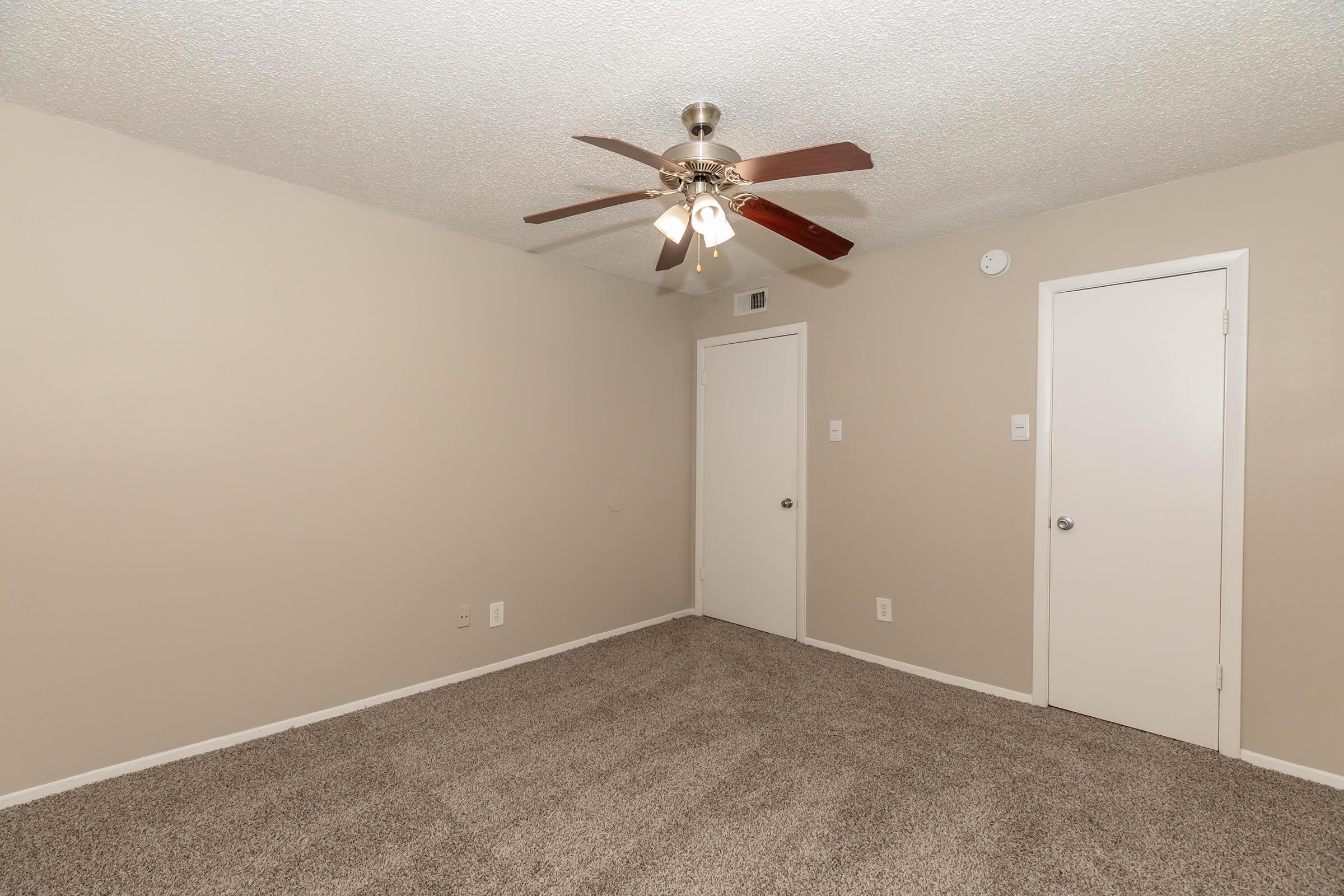 A spacious, empty room with beige walls and light brown carpeting. It features a ceiling fan with wooden blades, two doors (one leading to a closet and the other to an adjacent space), and a light fixture on the ceiling. The room is well-lit and has a simple, modern design.