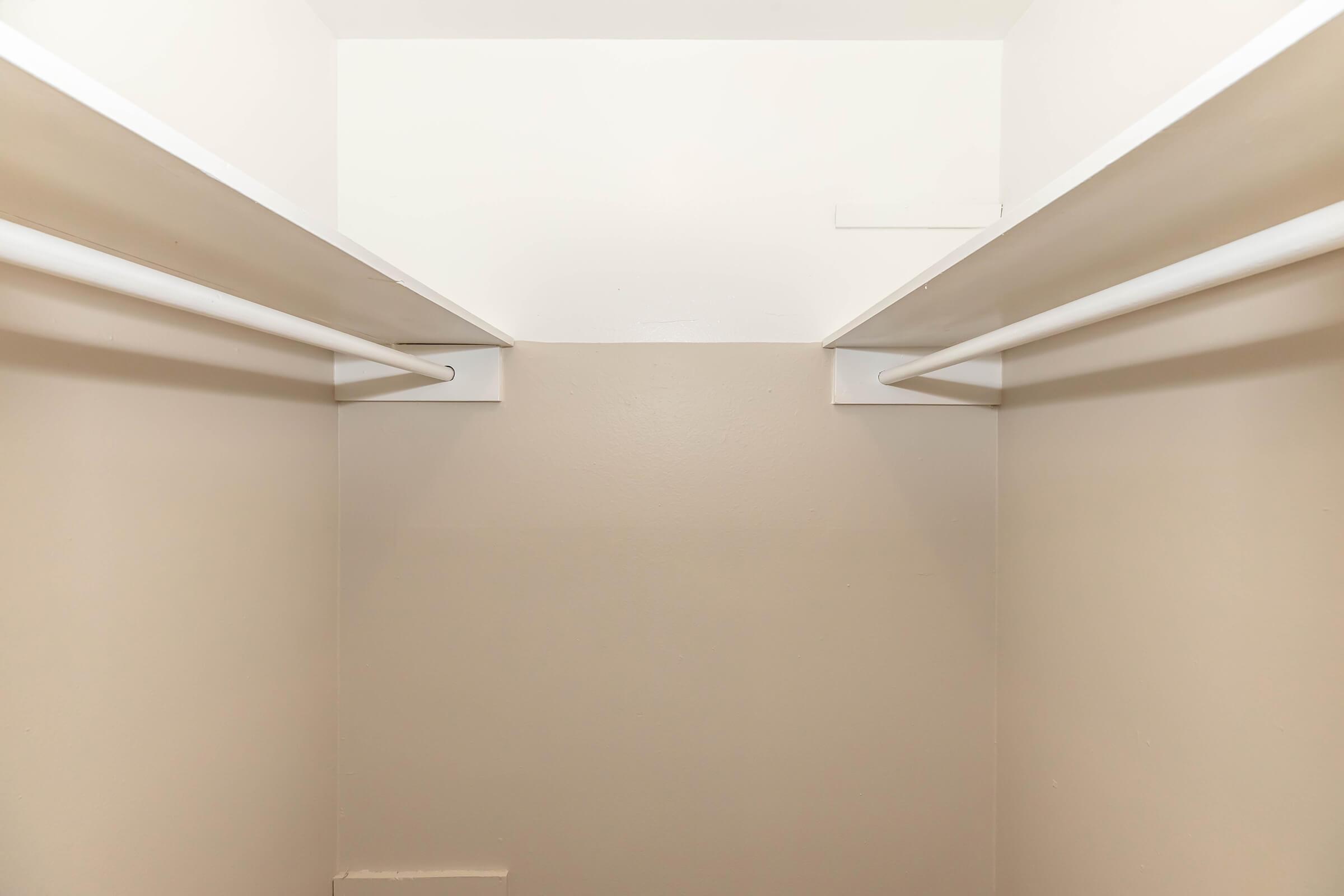 A view inside an empty closet, featuring two white horizontal rods for hanging clothes installed on opposite walls. The walls are painted a neutral beige color, and the ceiling is white. The space appears clean and organized, with no items stored inside.