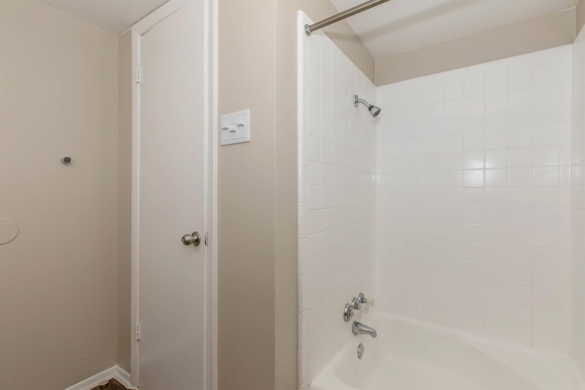 A small bathroom featuring a shower area with white tiled walls, a bathtub, and a closed door. The space has neutral-colored walls and a light floor, creating a clean and simple aesthetic. A showerhead and faucet are visible, but there are no decorative elements present.