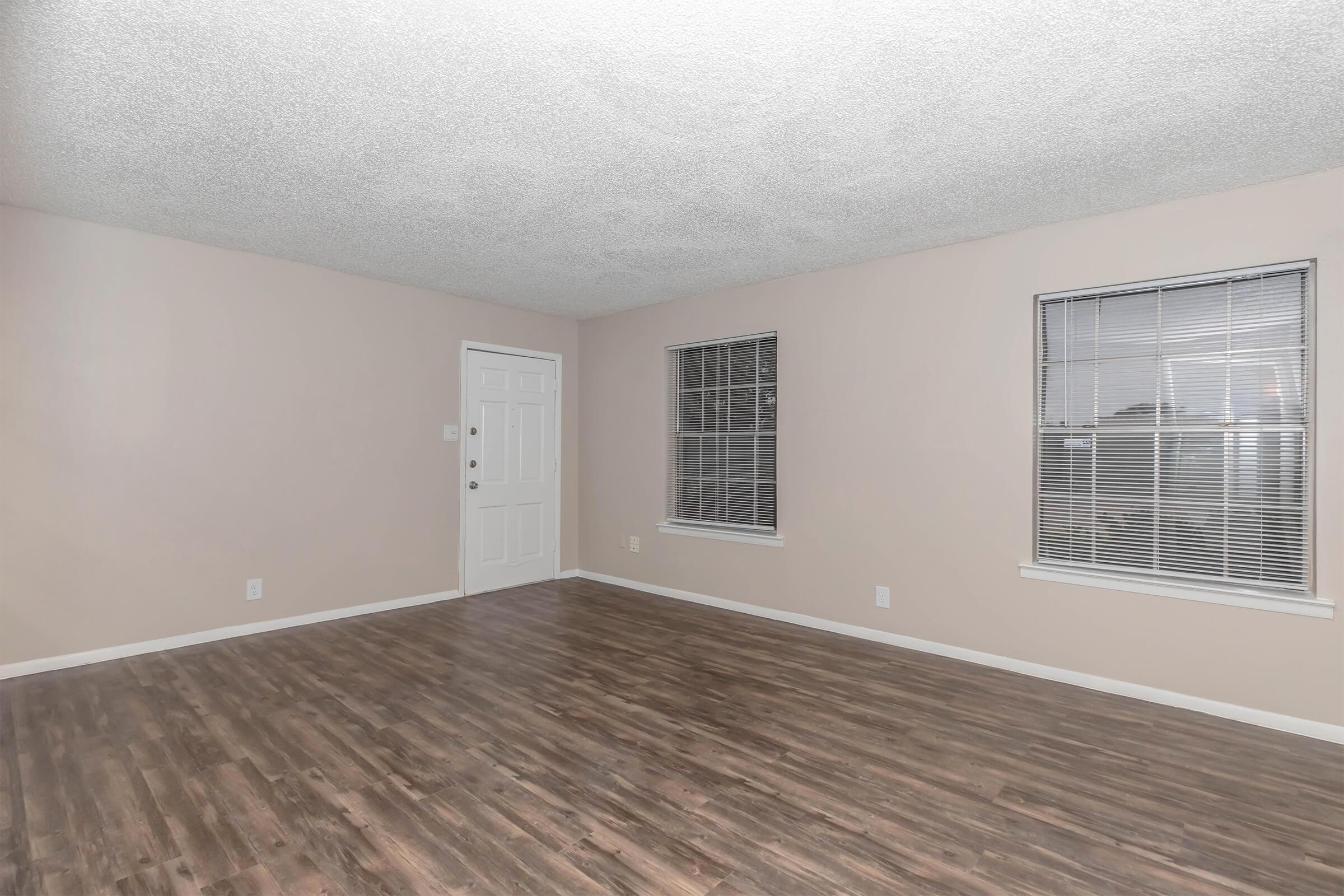 An empty room with light beige walls and a textured ceiling. It features two windows with blinds, a white door, and a floor made of dark wooden planks. The space is well-lit, appearing spacious and ready for furnishing.