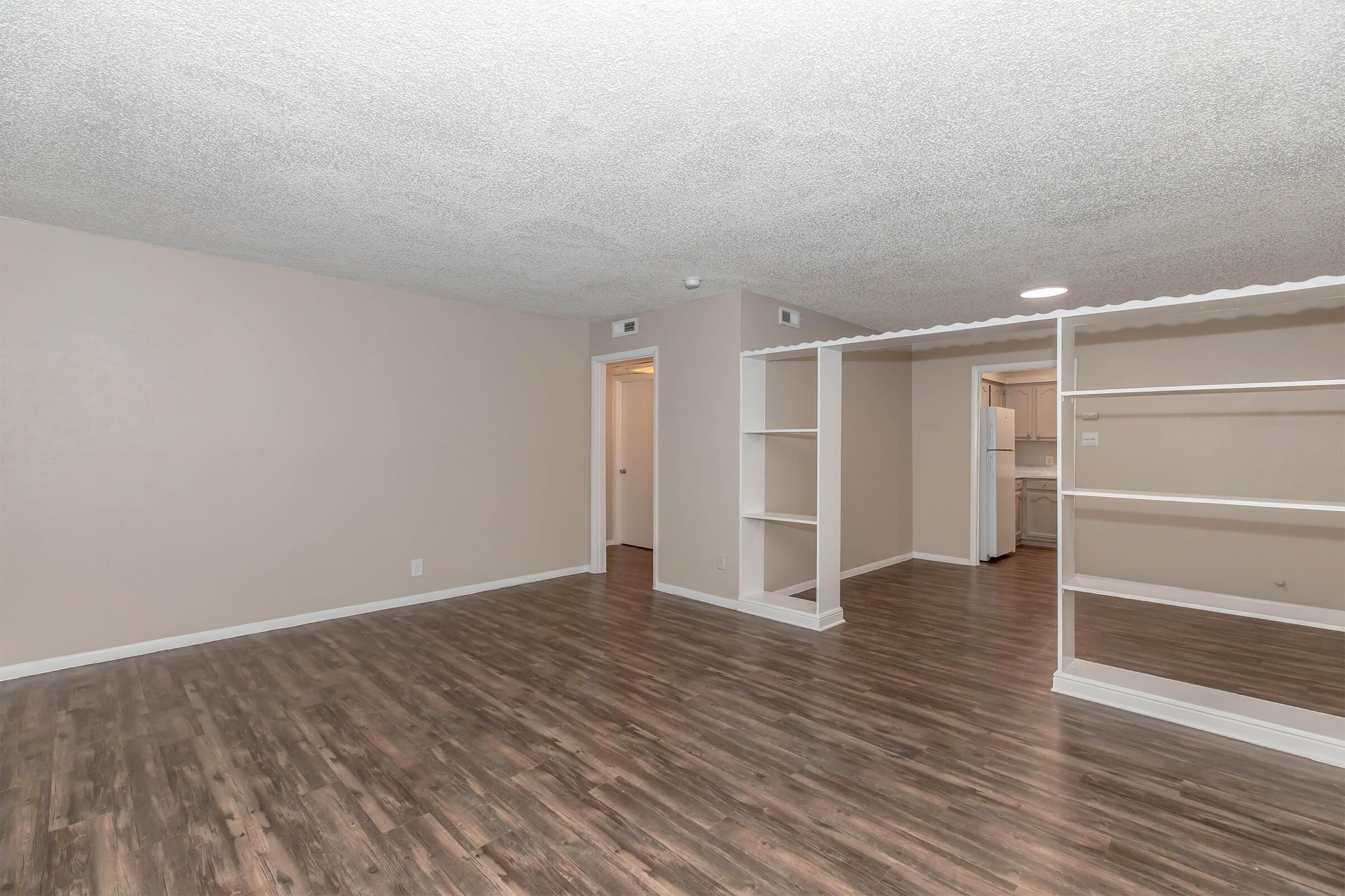 Spacious living area with a textured ceiling and laminate wood flooring. Light-colored walls and an open layout leading to a kitchen area with a refrigerator visible in the background. A narrow shelf divides the space, enhancing the room's airy feel. Doorways lead to additional rooms.