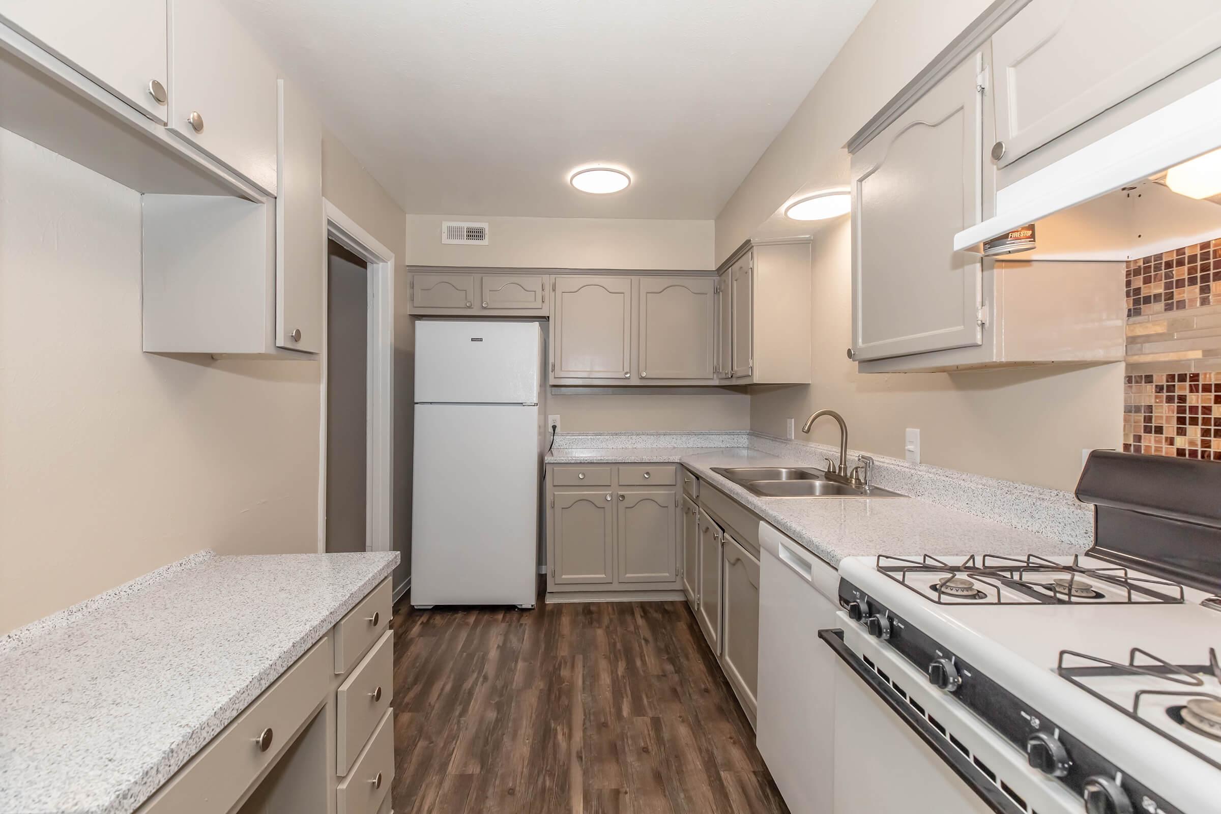 A modern kitchen featuring light gray cabinetry, a white refrigerator, and a stove with a grill. The countertops are light-colored, and there's a tiled backsplash. The floor is a dark wood finish, creating a contemporary and clean aesthetic with ample storage space.