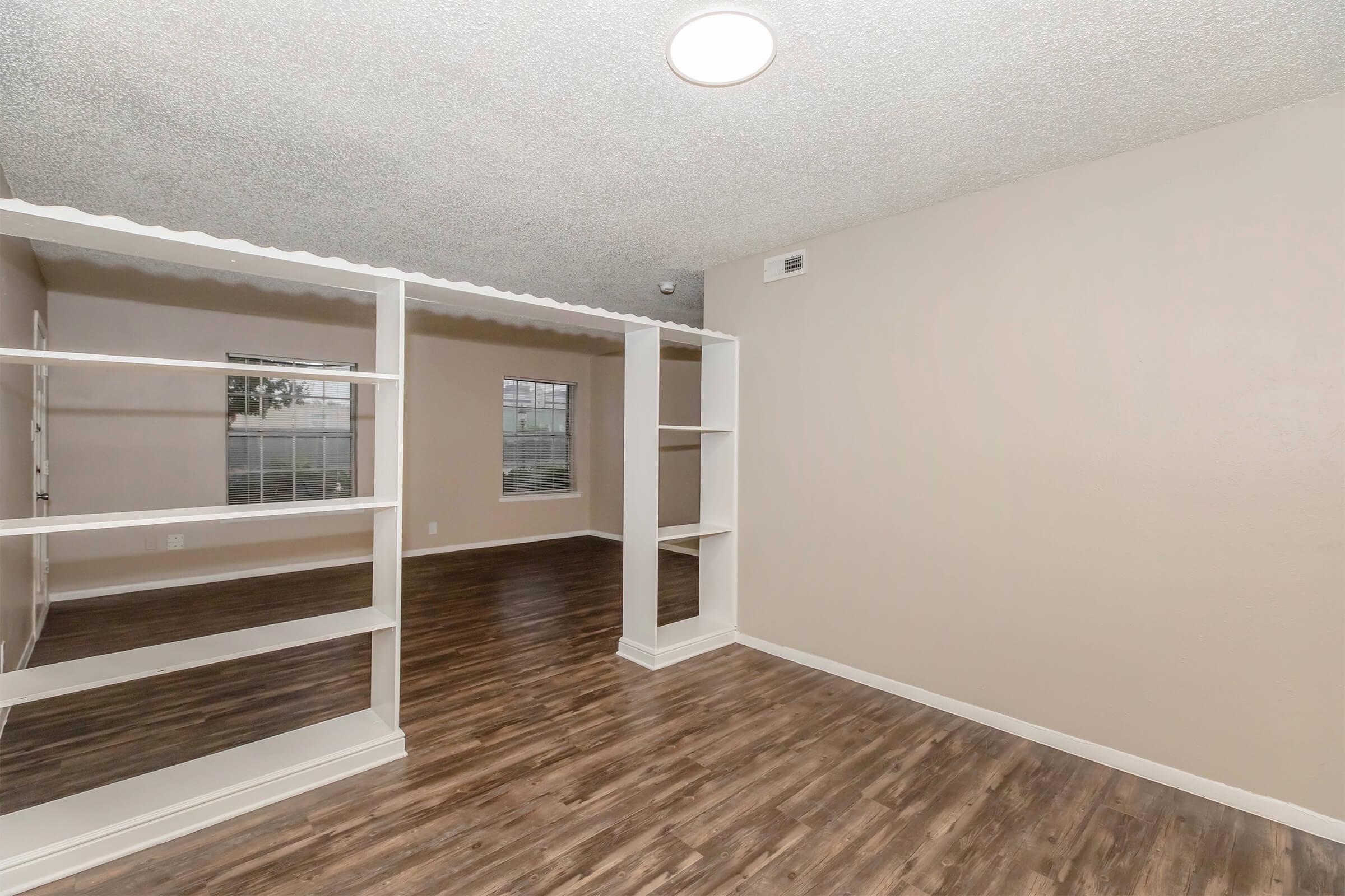 A spacious, empty room featuring light brown walls, laminate flooring, and a ceiling light. There are built-in white shelves separating areas, and two windows with views of the outside. The room is designed for versatile use, providing ample natural light.