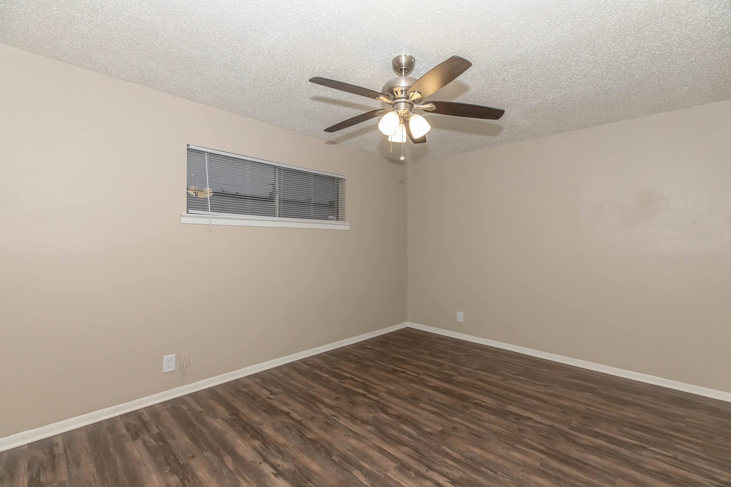 Empty room featuring a ceiling fan with four light fixtures, a window with blinds, and wooden flooring. The walls are painted a soft beige color, and there are no furnishings or decorations, creating a clean and minimalist aesthetic.