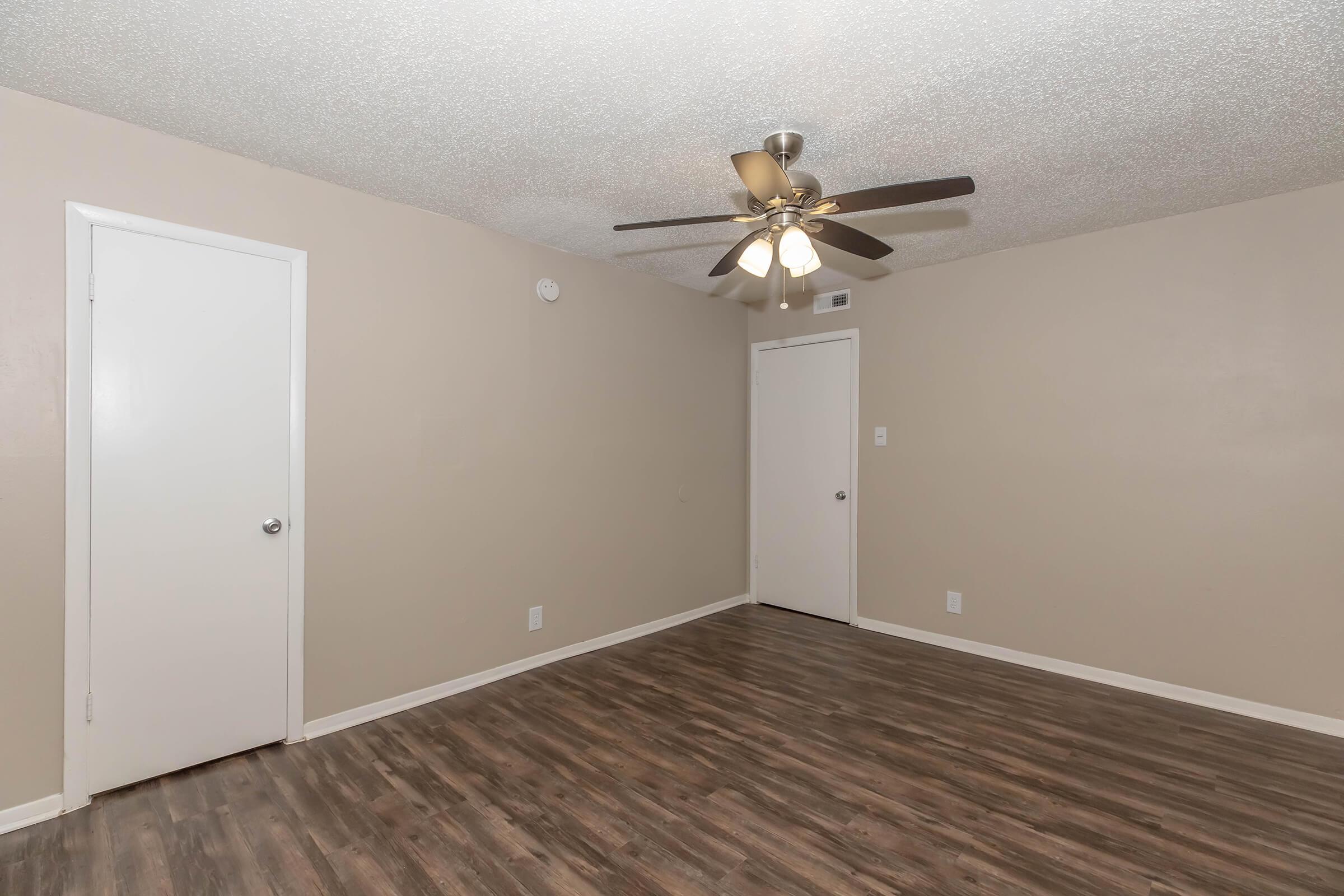 A small, light-colored room with a textured ceiling and a ceiling fan. The walls are painted beige, and there are two white doors—one visible on the left and the other on the right. The floor is wooden laminate, giving the space a clean and modern appearance.