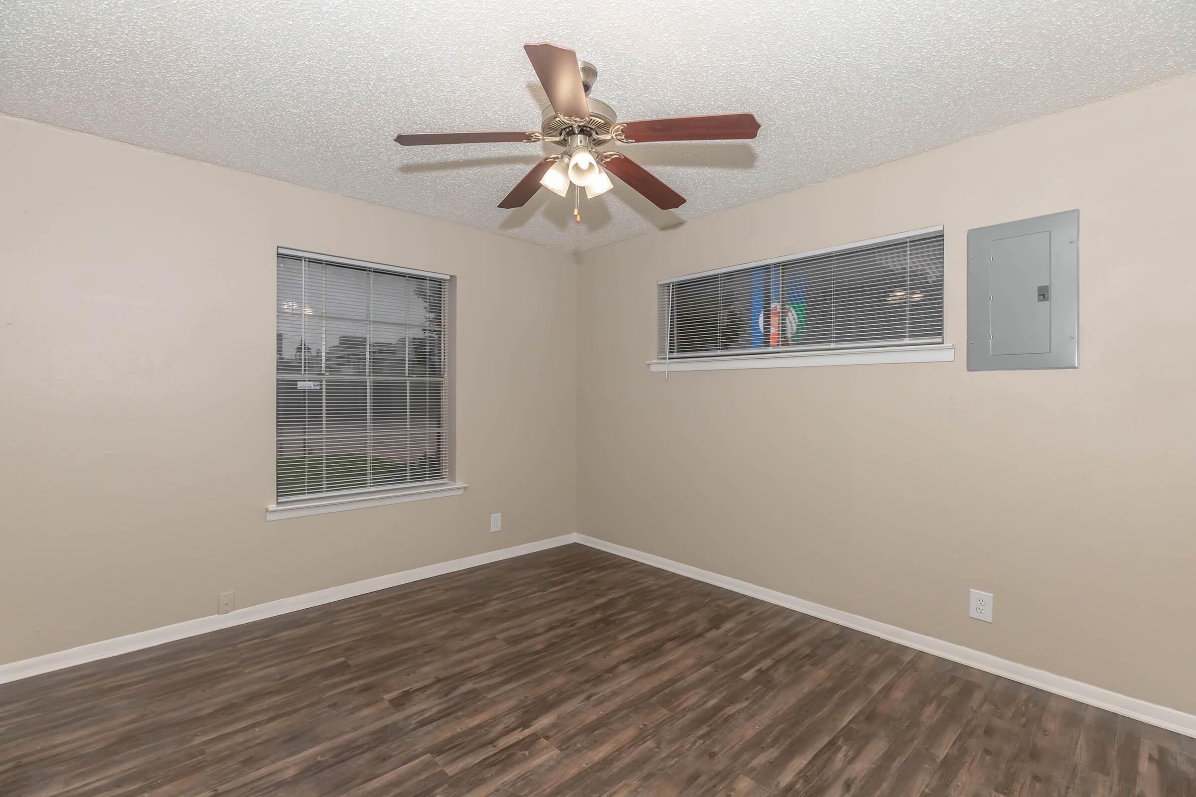 A well-lit, empty room featuring light-colored walls, a ceiling fan with light fixtures, two windows with blinds, and a brown laminate floor. An electrical panel is mounted on one wall. The space is ready for furniture and decoration.