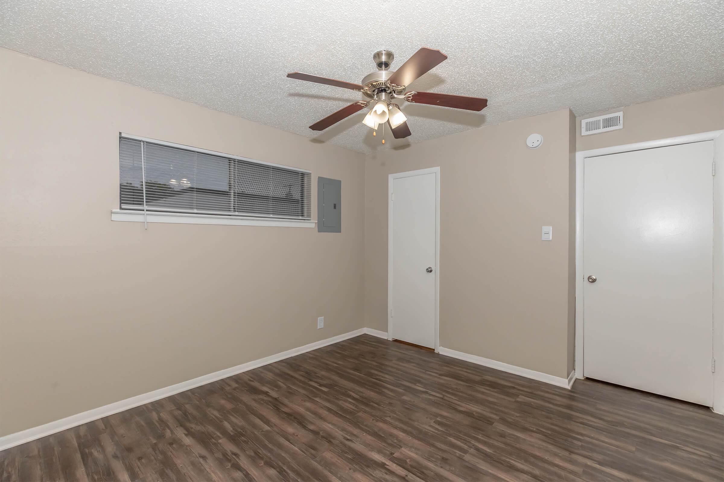 A spacious room featuring a light brown ceiling and wall color, a ceiling fan with light fixtures, laminate flooring, a small window with blinds, and two doors—one leading to another room and one for a closet. The lighting is bright, highlighting the clean and simple design of the space.