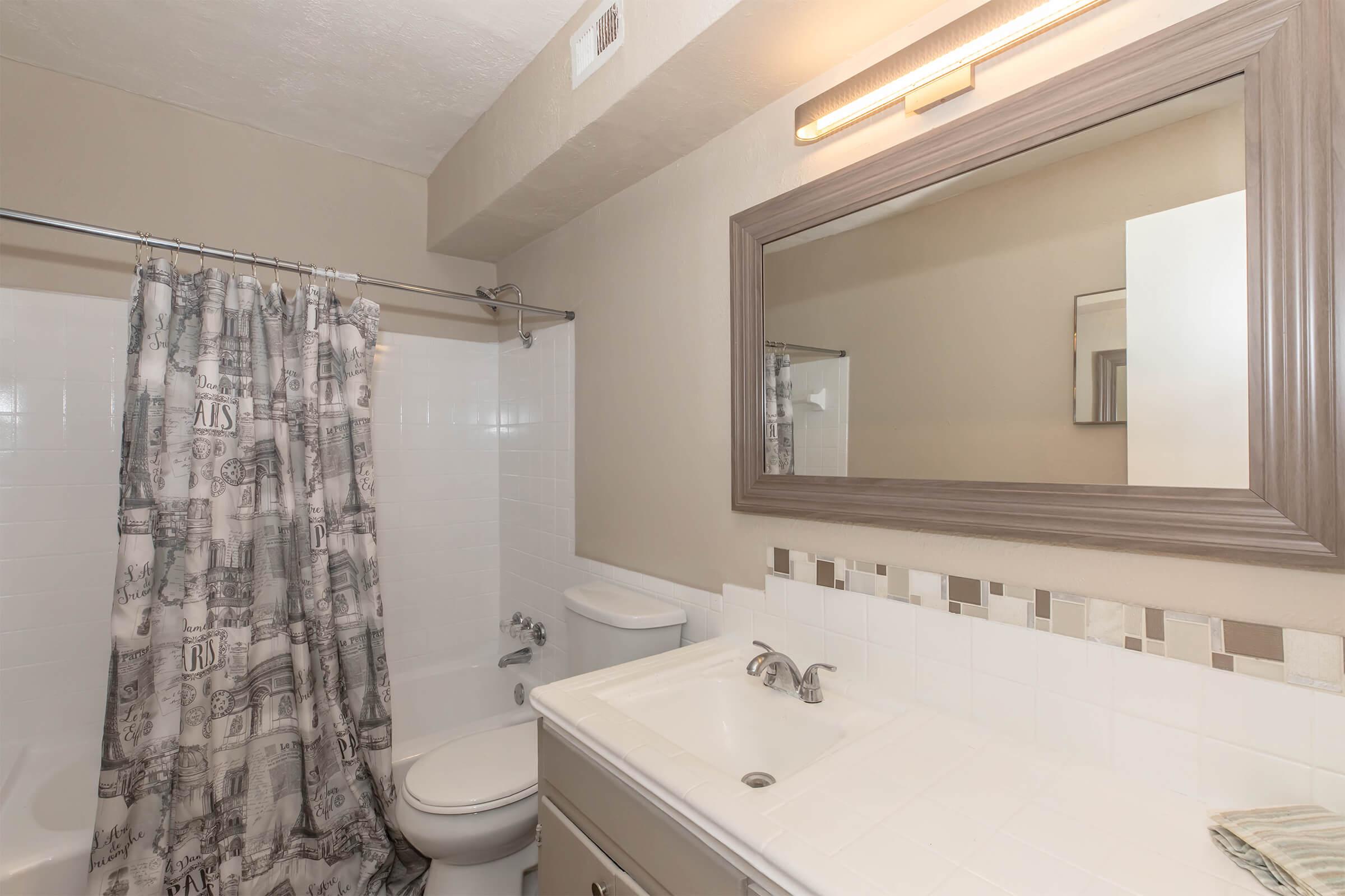 A modern bathroom featuring a shower with a patterned curtain, a white bathtub, a toilet, and a vanity with a sink. The walls are mostly white with a light taupe accent, and there's a large mirror above the sink. Soft lighting is provided by a lamp above the mirror.