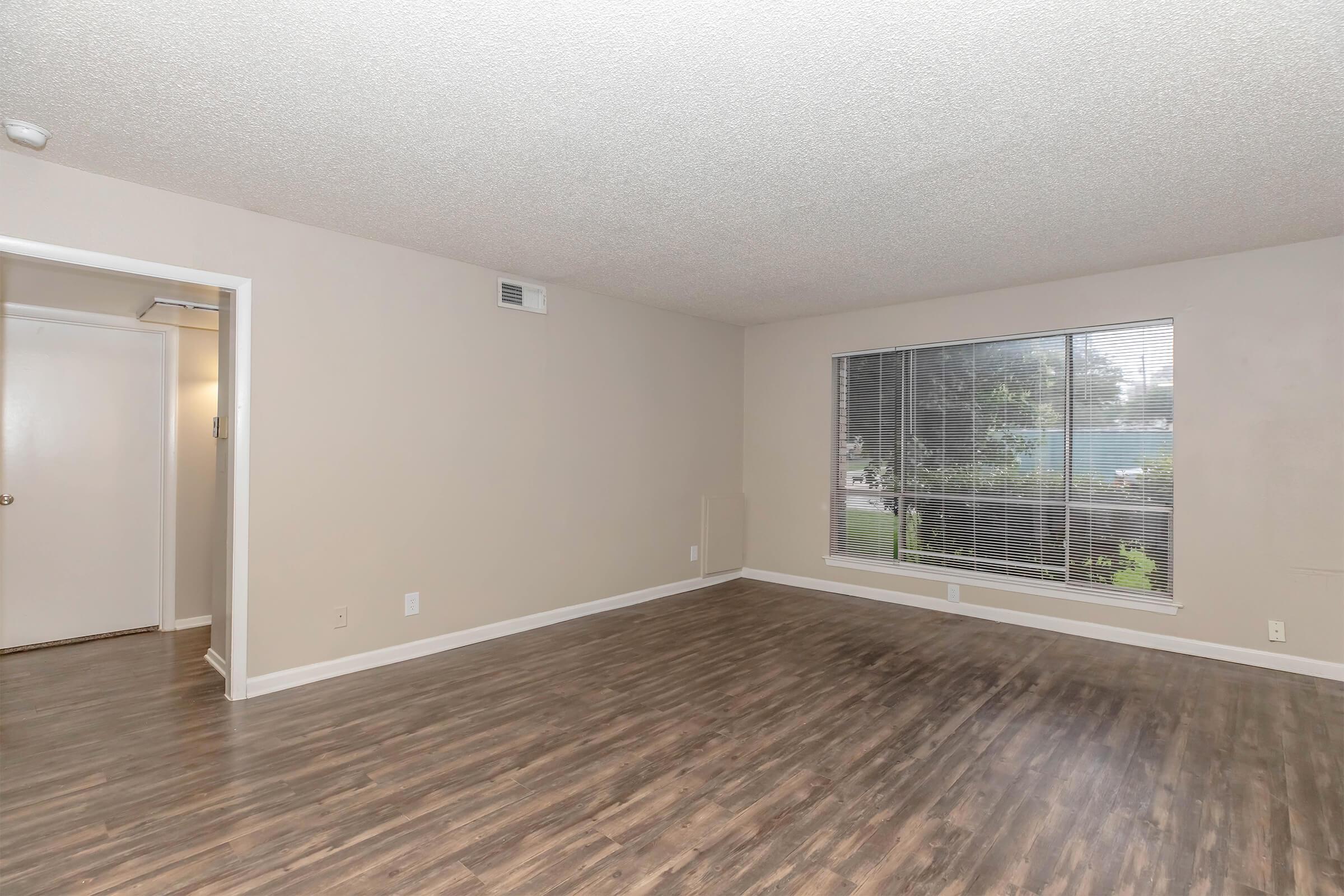 A spacious living room with light-colored walls and hardwood-style flooring. A large window allows natural light to fill the space, showcasing a view of greenery outside. An open doorway on the left leads to another room, creating an inviting and airy atmosphere.