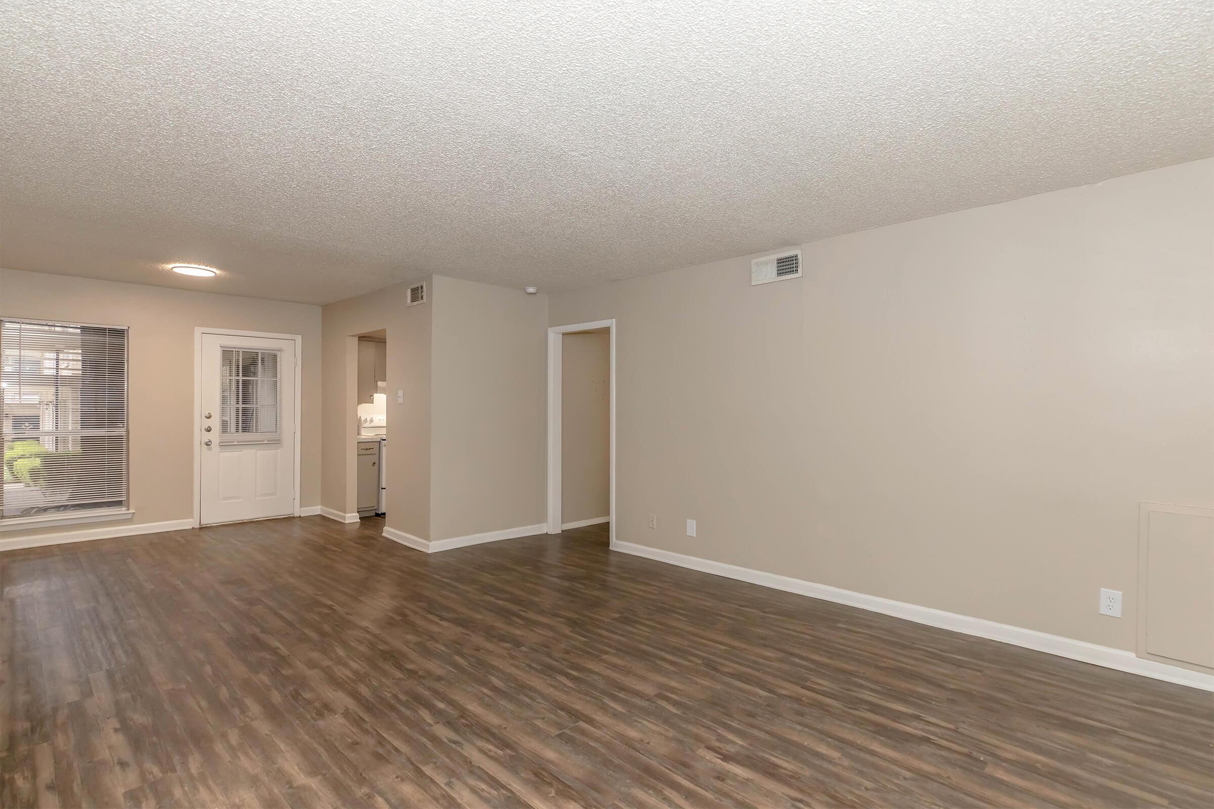 Spacious living area with light-colored walls and a textured ceiling. The room features laminate flooring, a front door with windows, and a doorway leading to a kitchen. A hallway to another room is visible, providing an airy and bright atmosphere.