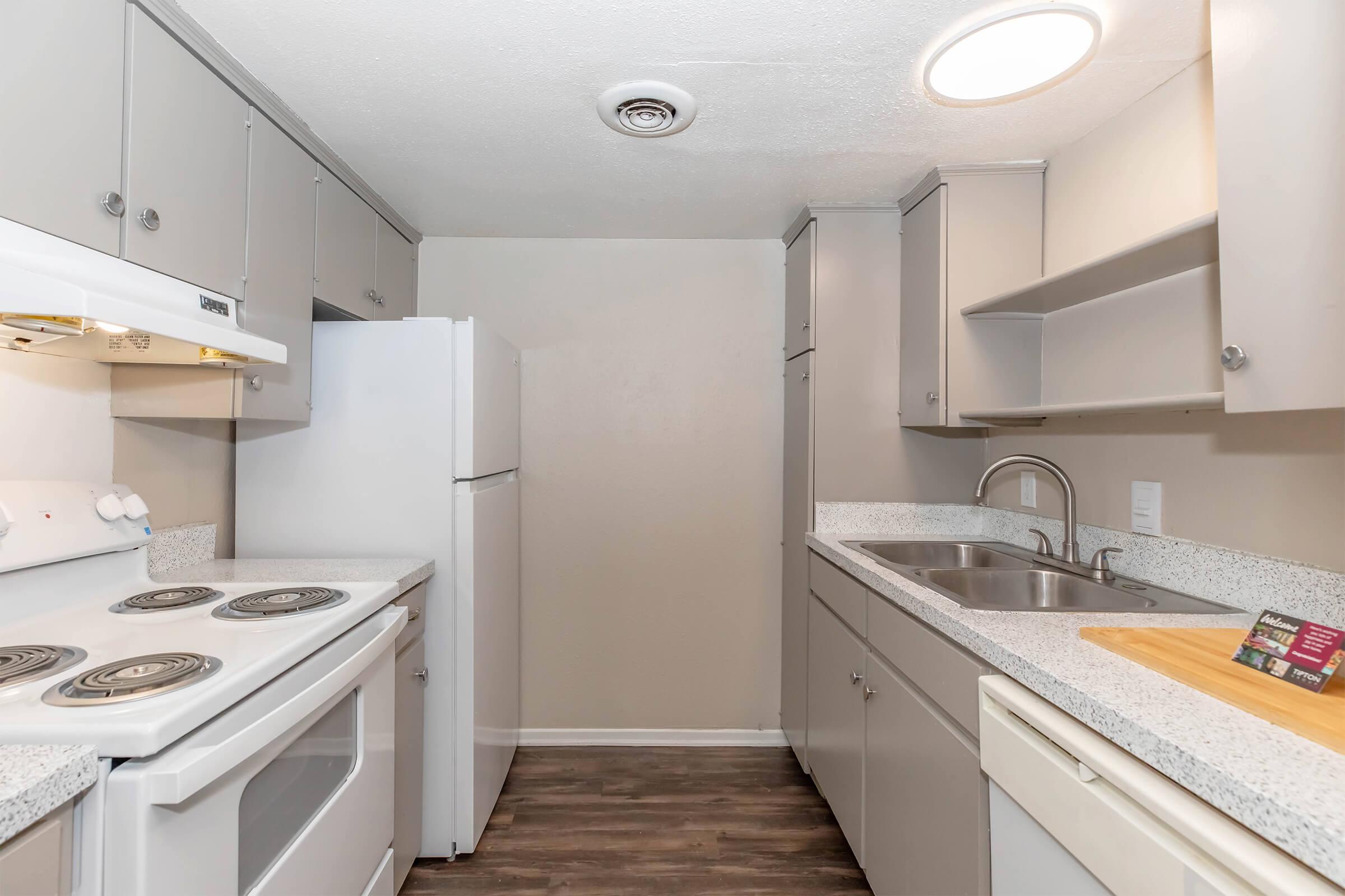 A compact kitchen featuring light gray cabinetry, a white stove, an oven, and a refrigerator. The countertop is gray with a matching sink and faucet. Natural light spills in from a ceiling fixture, enhancing the clean and modern design. The space is organized and functional, ideal for efficient cooking.
