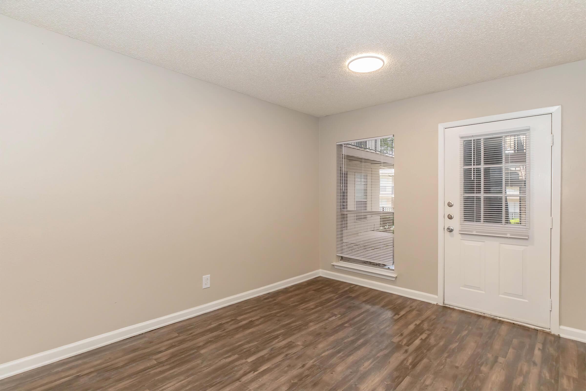 An empty room with light-colored walls and a textured ceiling. The floor is a dark, polished wood. There is a door on the right leading outside, and a window to the left with blinds, allowing natural light to enter. The space is uncluttered and ready for furniture.