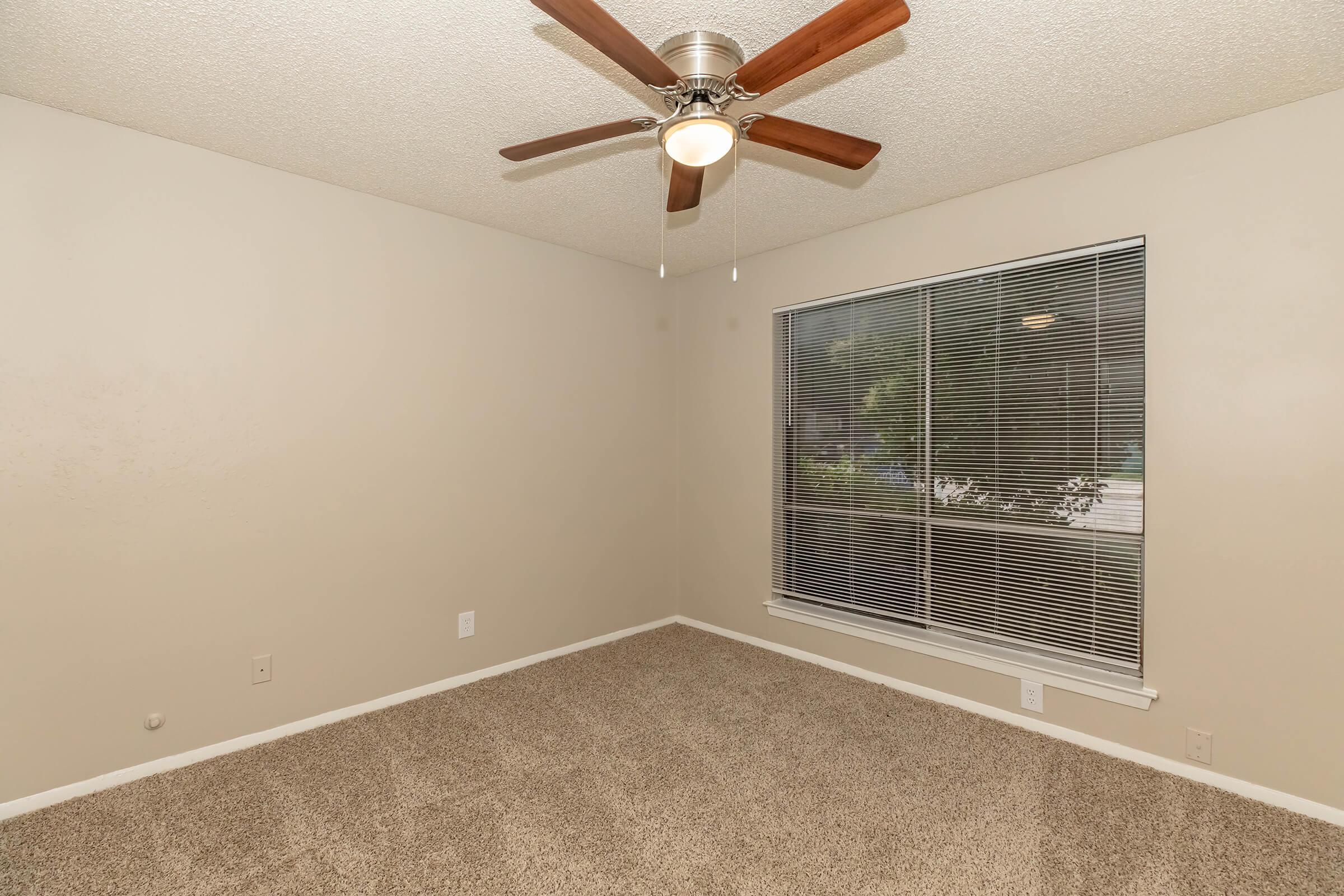 A small, empty room with beige walls and plush carpet. The ceiling features a modern ceiling fan with wooden blades. A large window with blinds allows natural light to enter, looking out to greenery. The space is unfurnished, ideal for personalization.