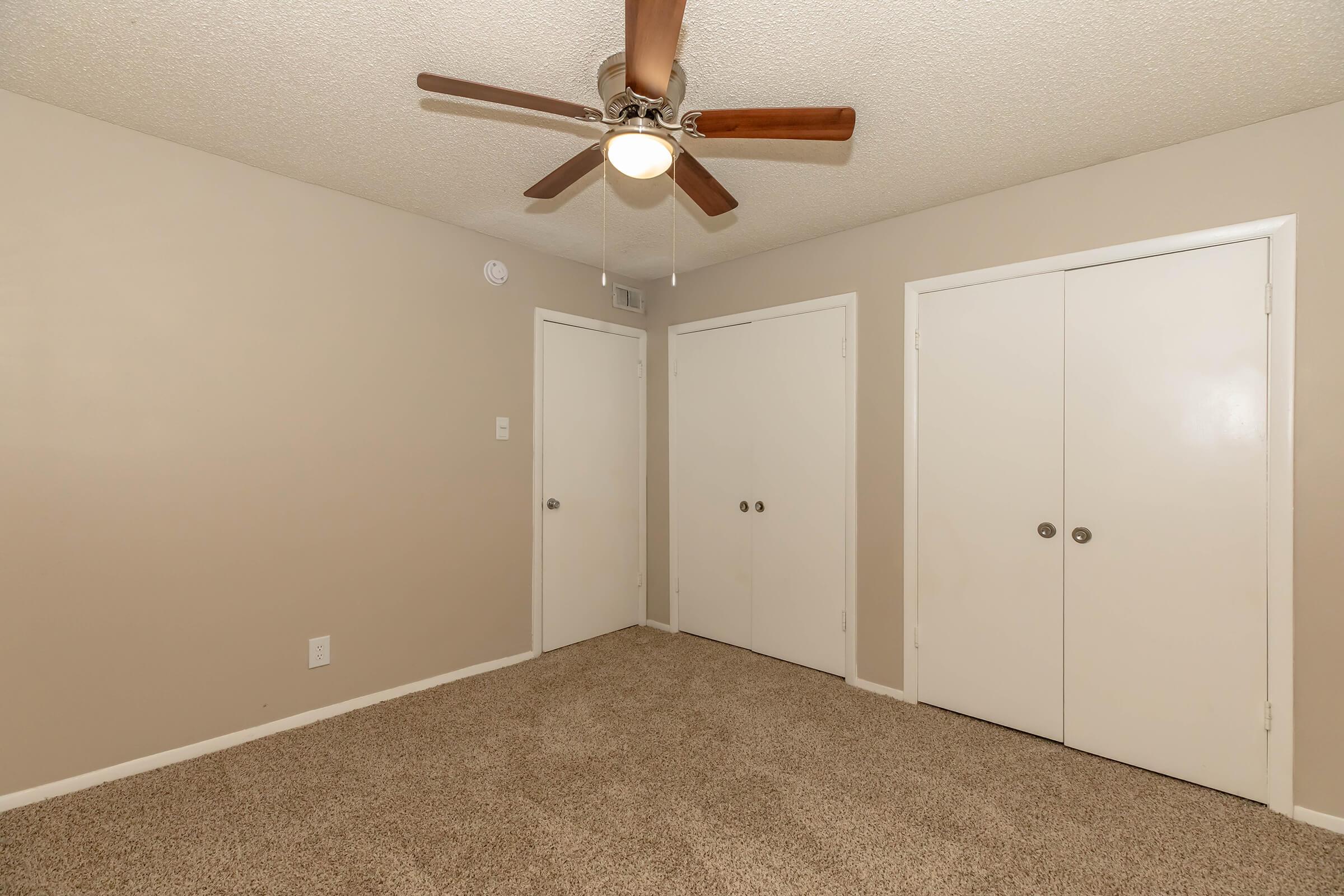 A small, empty room with light beige walls and a ceiling fan with wooden blades. The floor is covered in carpet, and there are two closed white closet doors on the right side. A single light fixture hangs from the ceiling, providing soft illumination. The room has a clean and simple appearance, suitable for various uses.