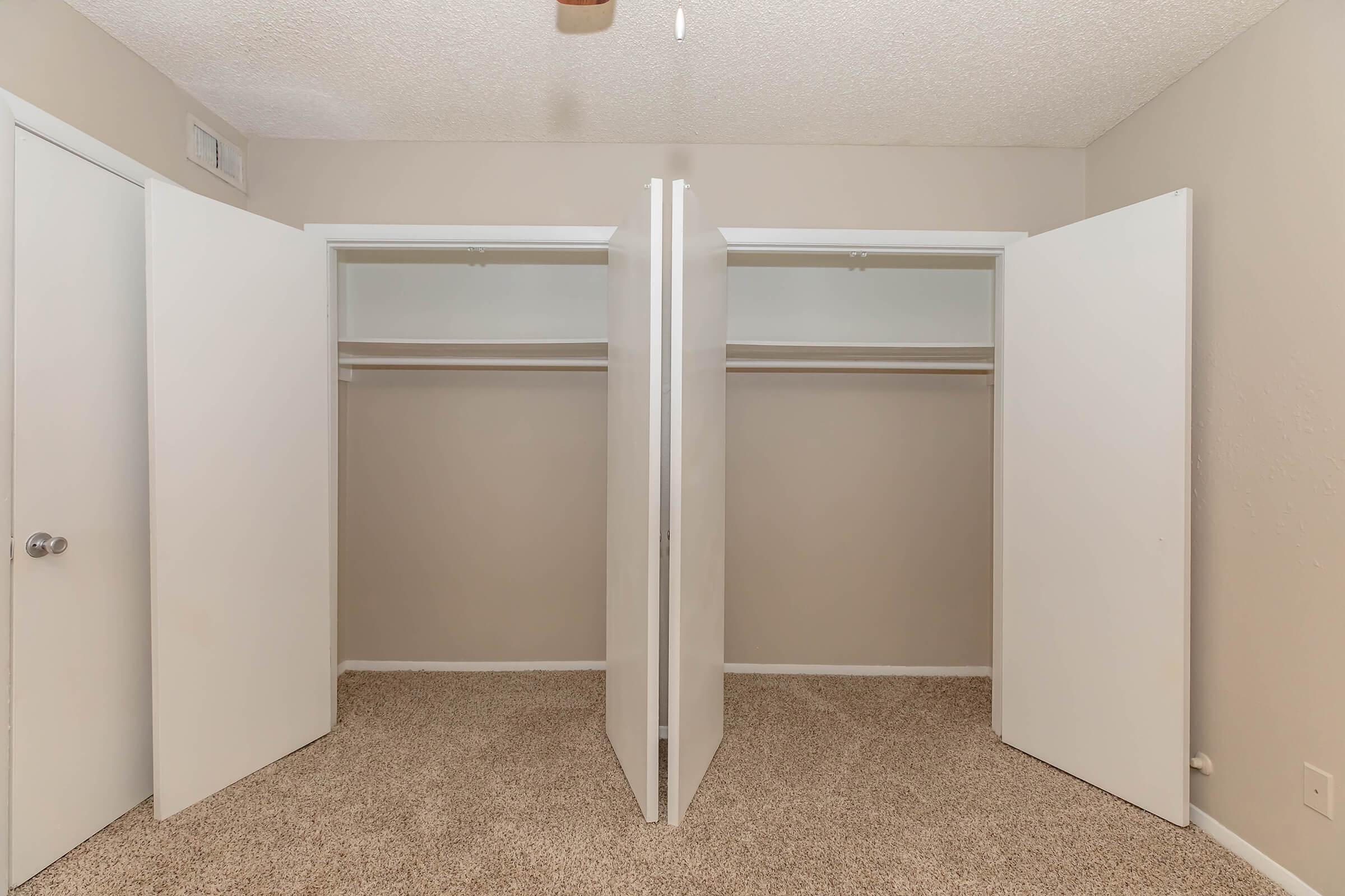 A well-lit room featuring two open white sliding closet doors, showcasing empty shelves and hanging rods. The walls are painted a light neutral color, and the floor is covered with beige carpet, creating a clean and spacious appearance.