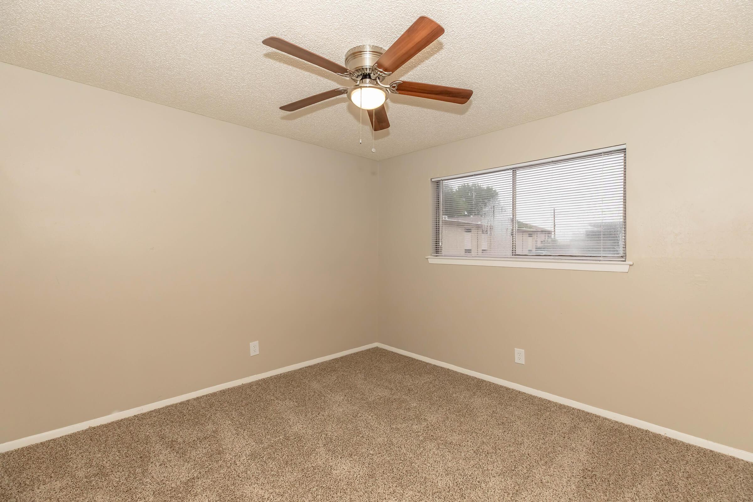 A neutral-toned empty room featuring a ceiling fan with wooden blades, a window with blinds allowing natural light, and beige carpet flooring. The walls are painted in a soft cream shade, giving the space a bright and inviting feel. Ideal for customization or decoration.
