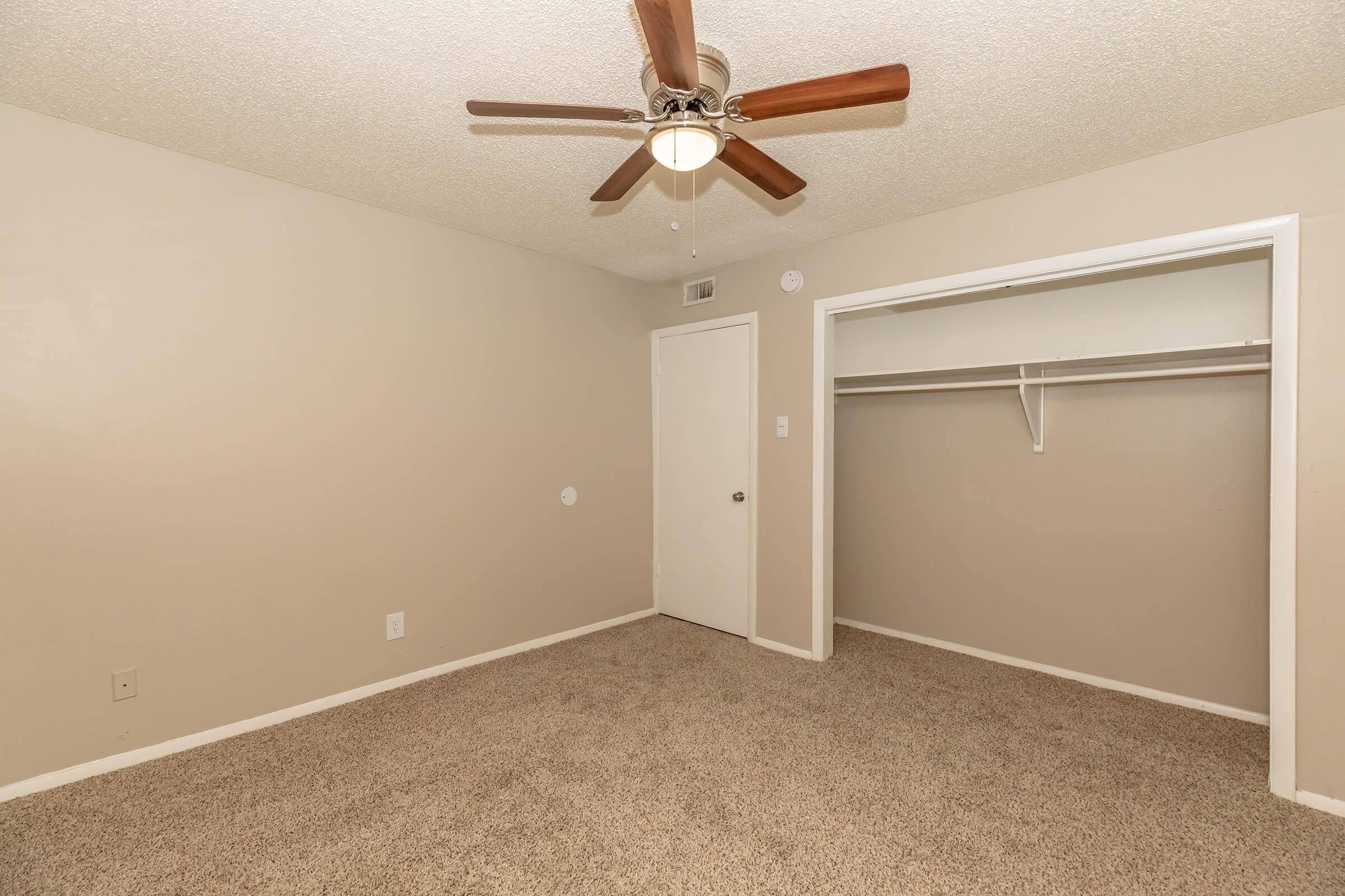 A spacious and neutral-colored bedroom featuring a ceiling fan, light-colored walls, and soft carpet. There is a closet with a single shelf and a door leading to another room. The room has a bright appearance, offering ample space for furniture placement.