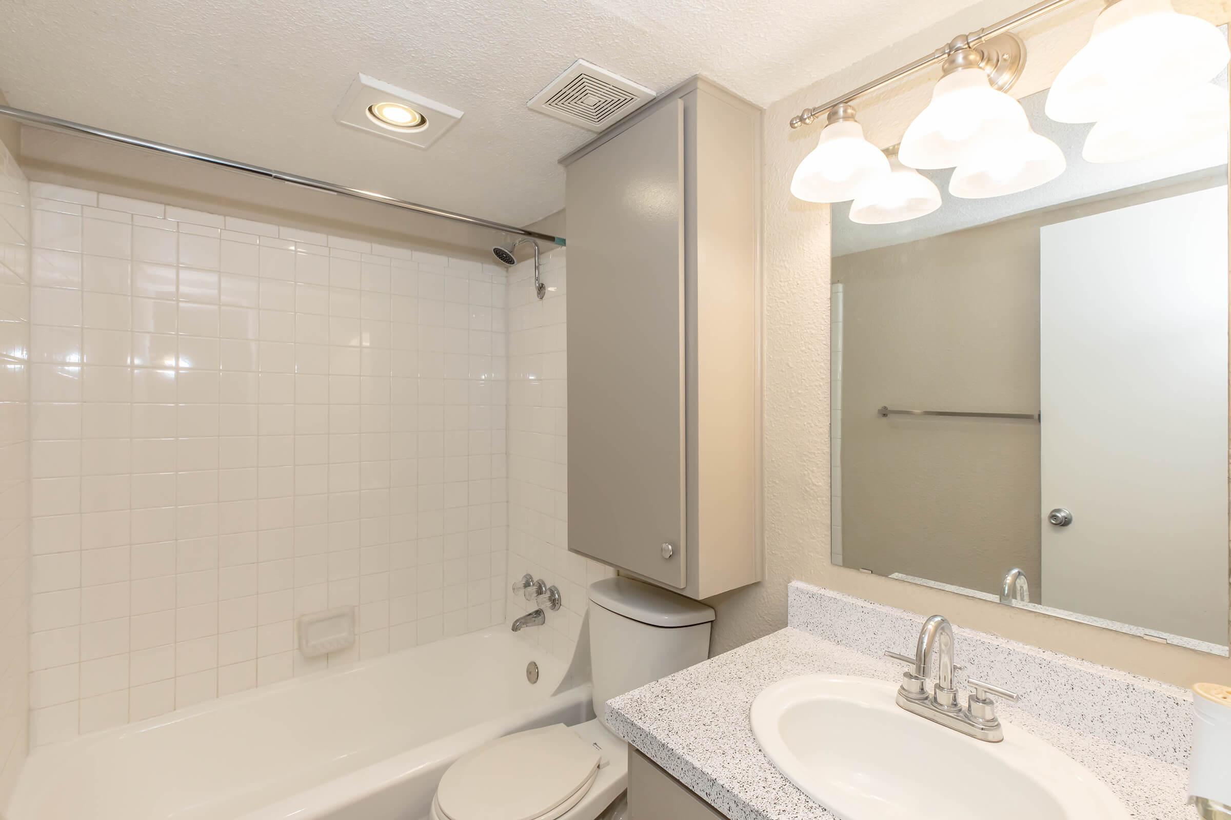 A clean, modern bathroom featuring a white tiled shower and bathtub combination, a toilet, and a granite countertop with a sink. The space is well-lit by three frosted glass light fixtures above the mirror, and there is a neutral color scheme throughout.