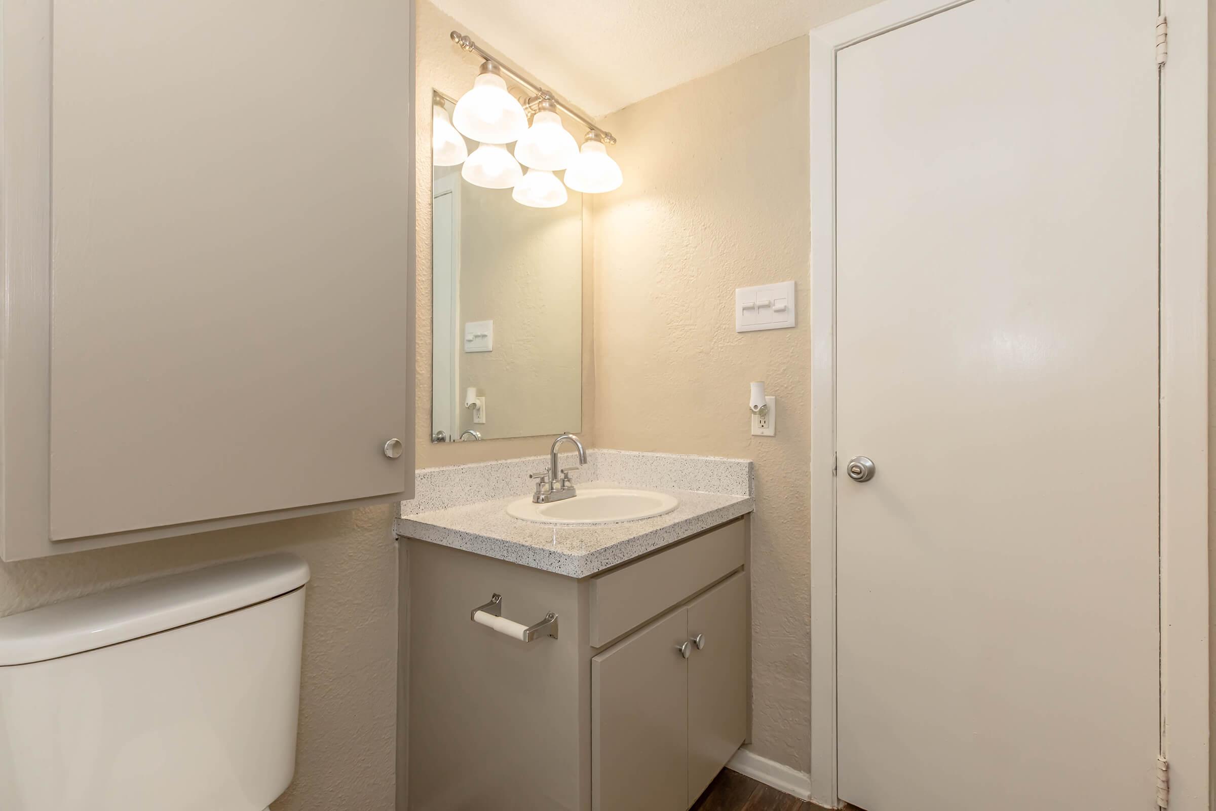 A clean, modern bathroom featuring a light-colored countertop with a sink, mirrored light fixture above, gray cabinetry for storage, and a white toilet. The walls are painted a soft beige, and a closed door is visible on the right side of the image.