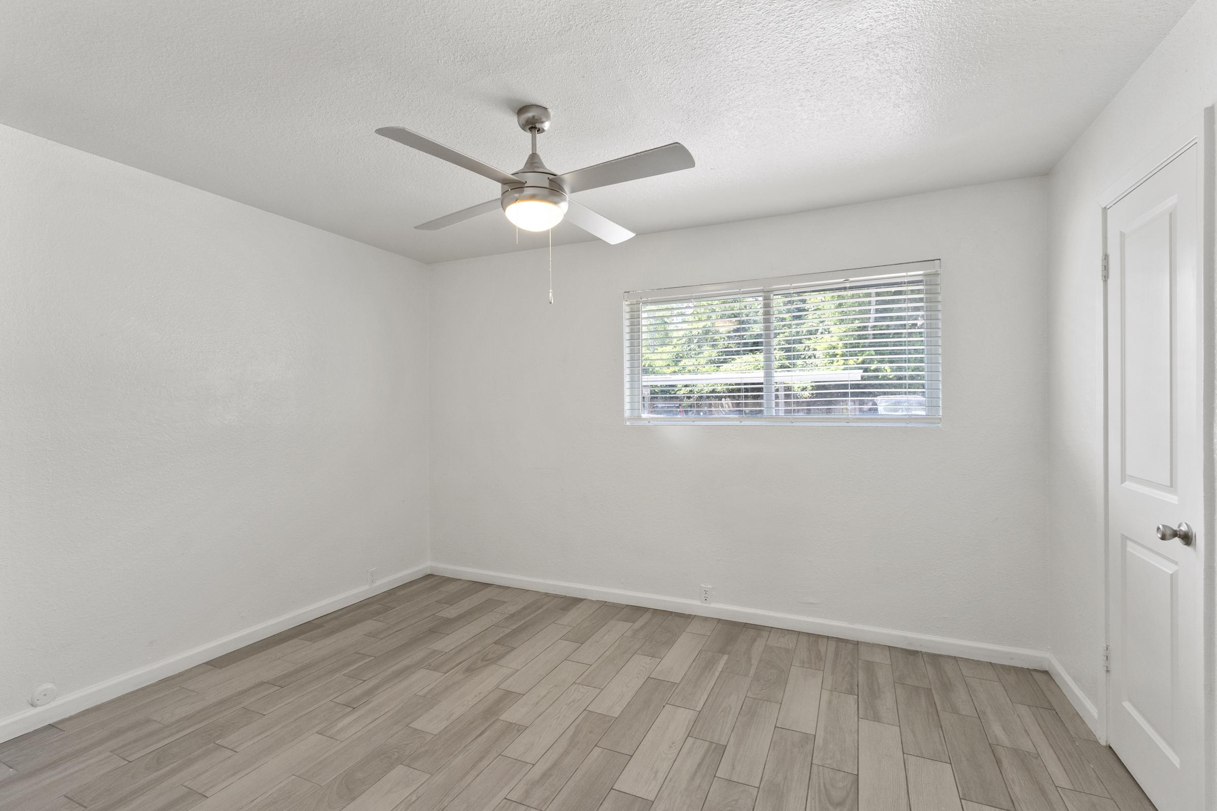 A bright, empty room with light-colored walls and tiled flooring. A ceiling fan is mounted in the center, and a window with blinds allows natural light to enter. The space is minimalistic, featuring no furniture, creating an open and airy feel.