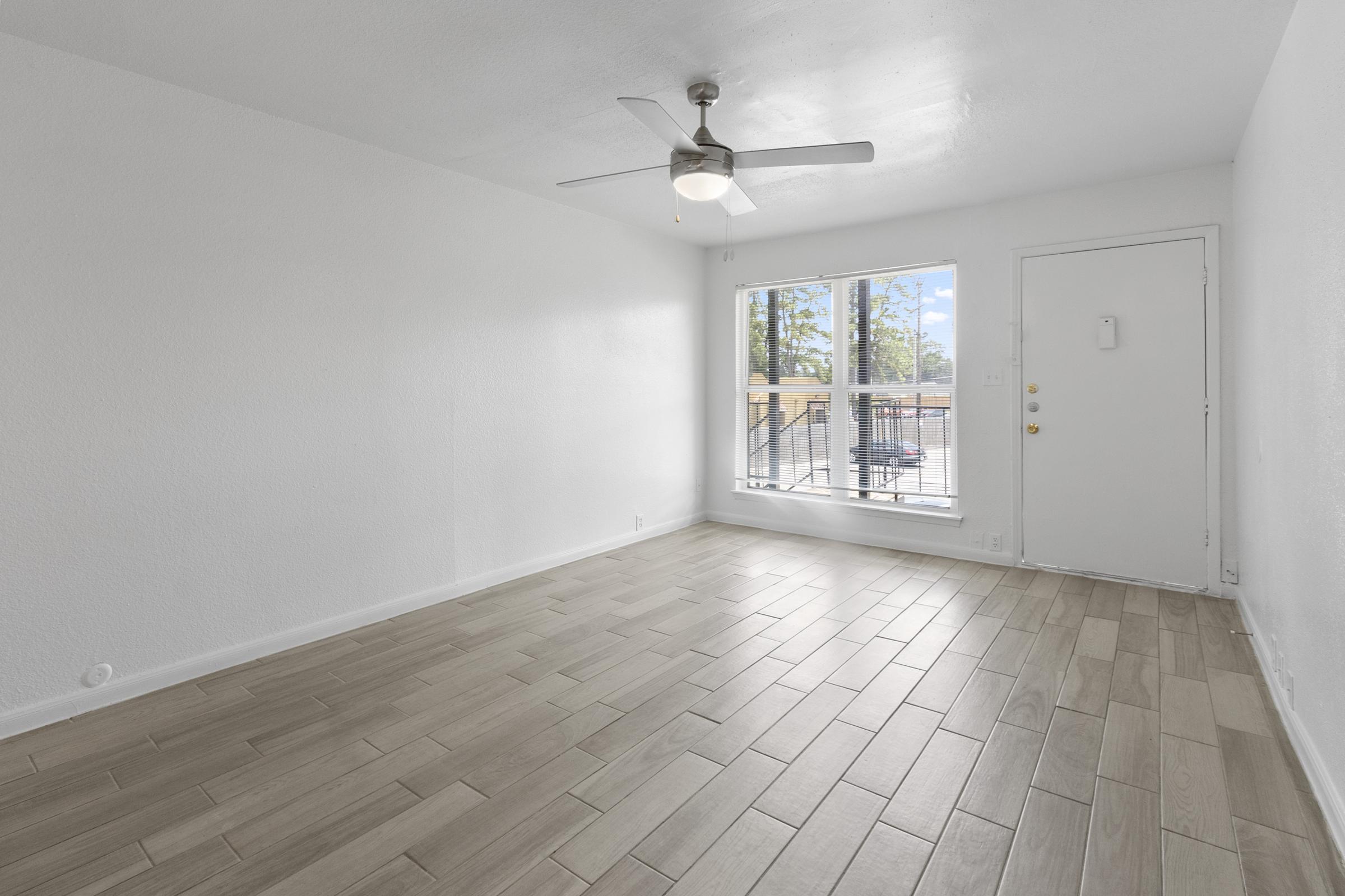 A spacious, bright living area with light-colored walls and tiled flooring. The room features a ceiling fan and a large window allowing natural light to enter. A door leads to the outside, and there is minimal furniture present, creating an open and airy atmosphere.