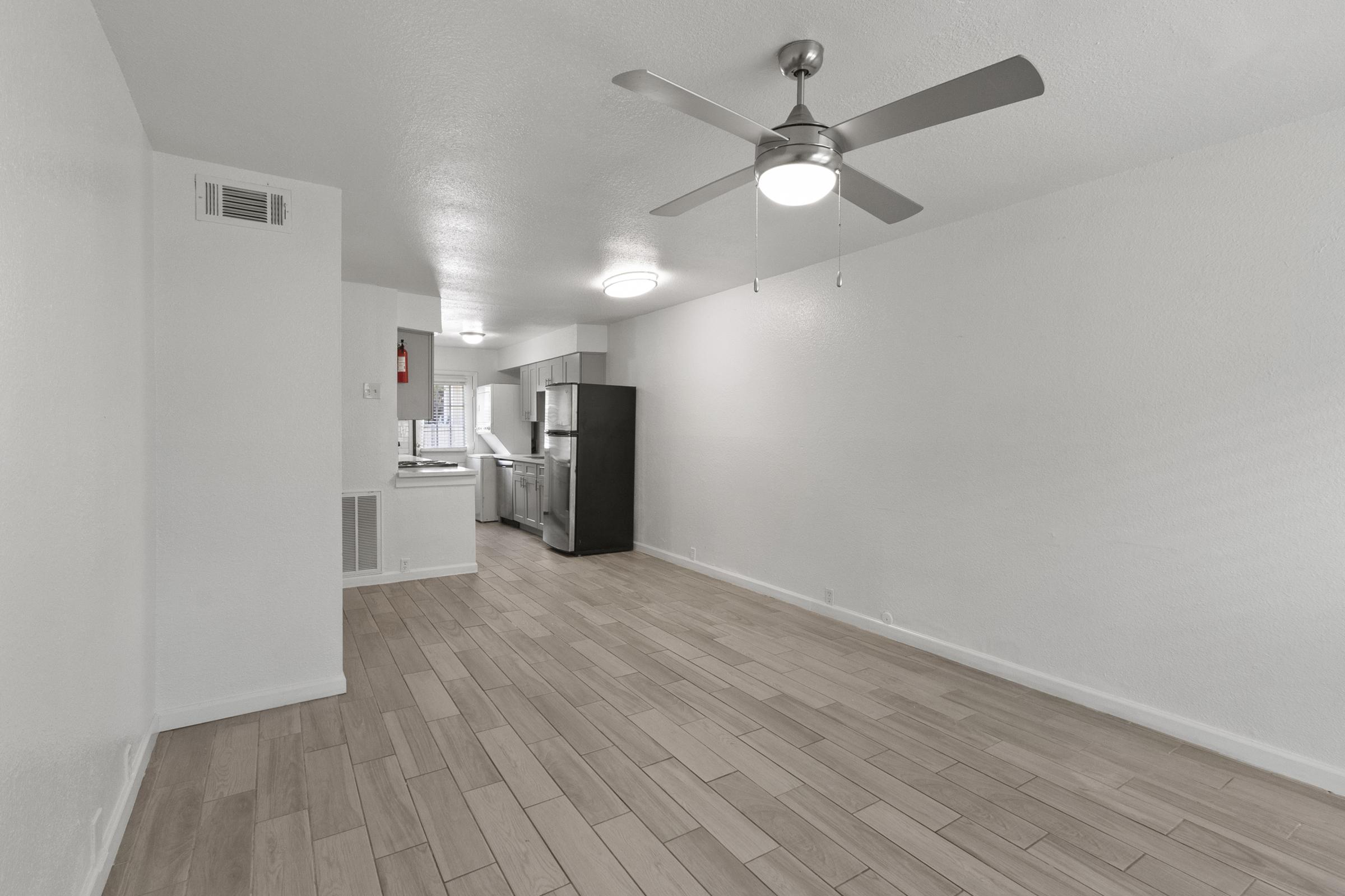 Interior view of a modern apartment featuring an open living space with light-colored wood flooring, a ceiling fan, and a window. The kitchen is visible in the background with stainless steel appliances and a bright ambience. The walls are painted white, contributing to a spacious feel.