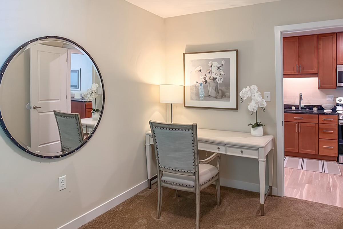 A well-decorated room featuring a round mirror, a stylish desk with a chair, a lamp, and a framed artwork of flowers. In the background, there is a small kitchenette with wooden cabinets, creating a cozy and inviting atmosphere.