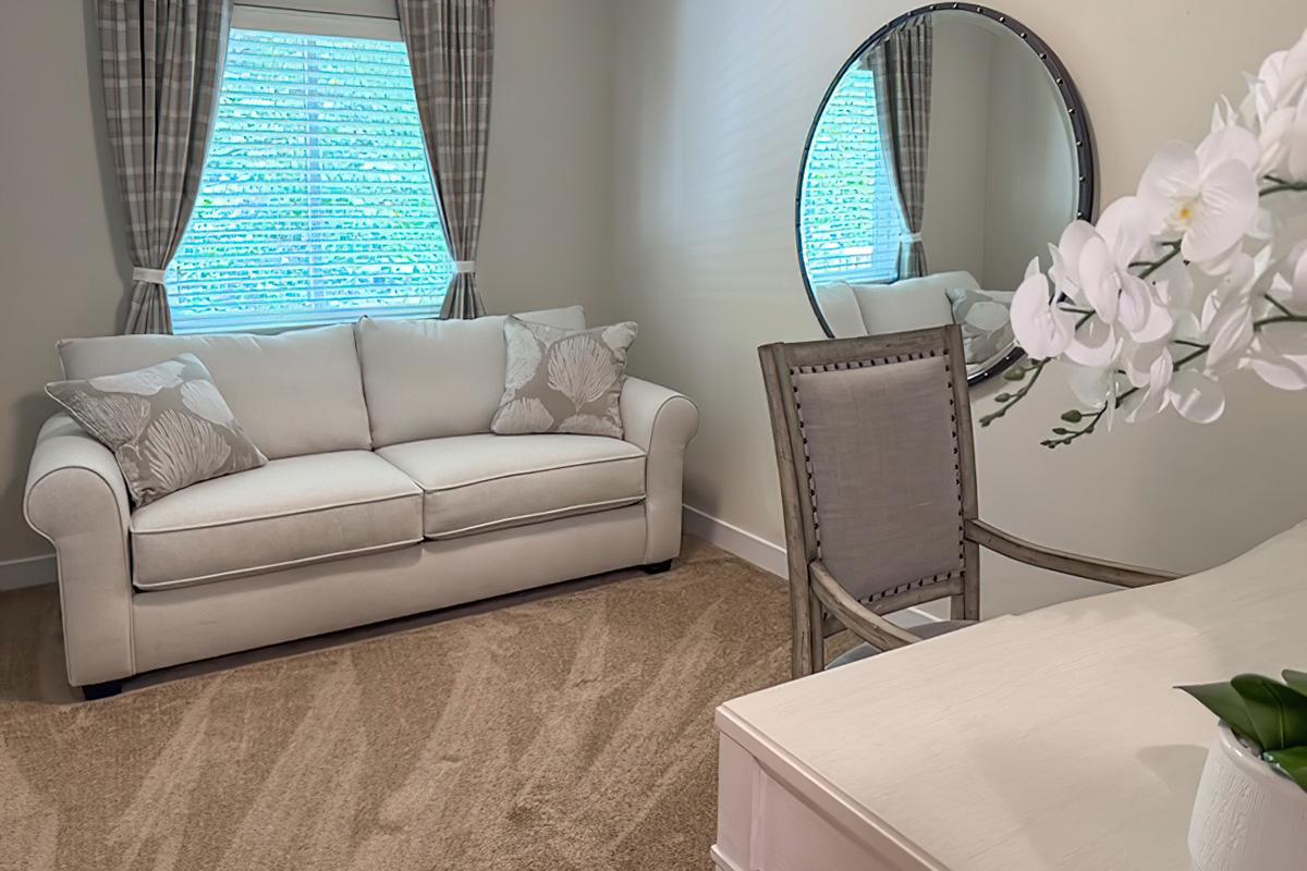 Cozy room featuring a light-colored sofa with decorative pillows, a round mirror on the wall, and a wooden chair next to a desk. Natural light filters in through a window dressed with checked curtains, while a vase of white orchids adds a touch of elegance to the space. Soft, neutral-toned carpet enhances the comfort of the room.
