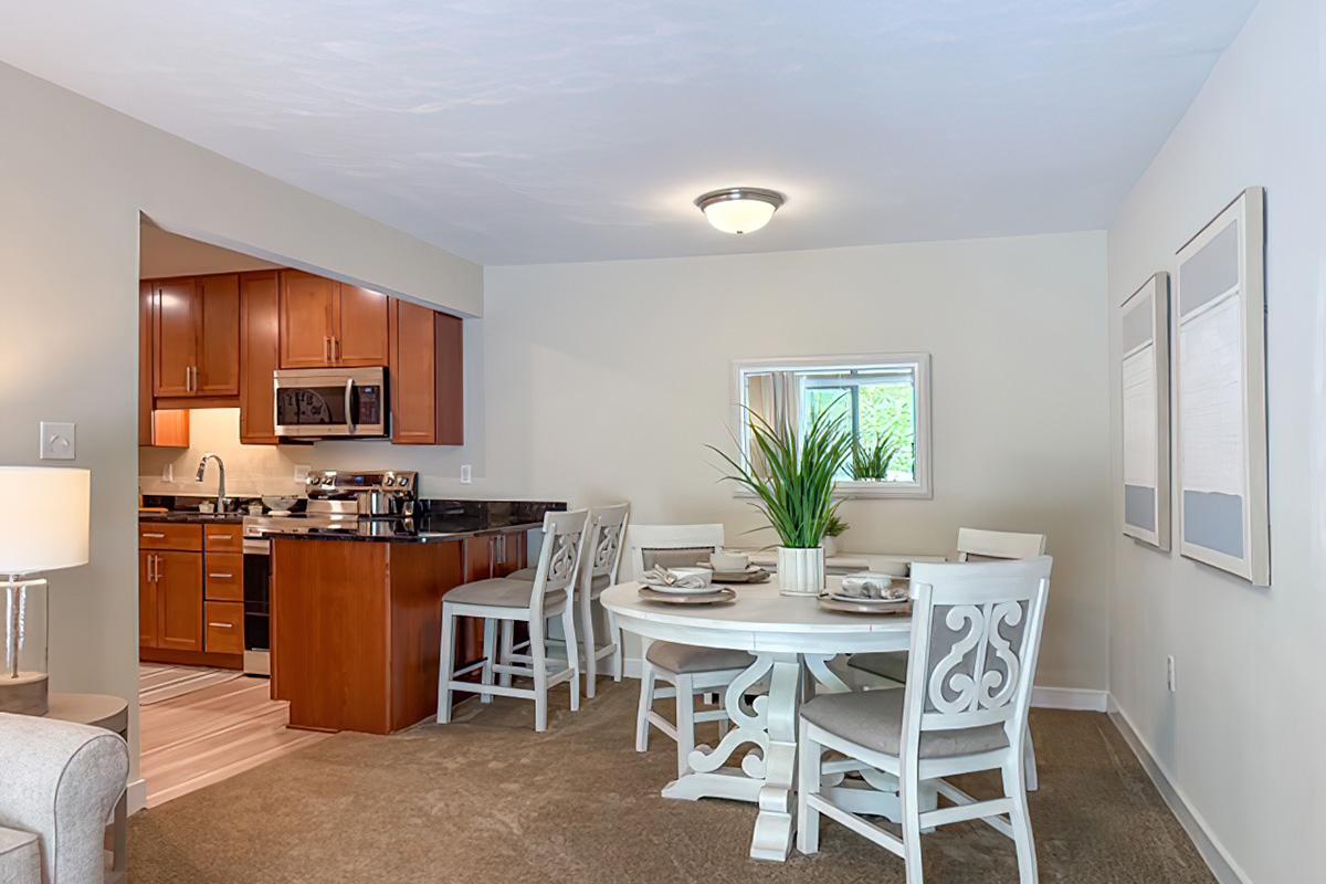 A cozy dining area featuring a round white table with decorative plates and a green plant centerpiece. The space is illuminated by a ceiling light, with a view into a modern kitchen that has dark wood cabinets and stainless steel appliances. The walls are painted in a soft neutral color, enhancing the warm ambiance.