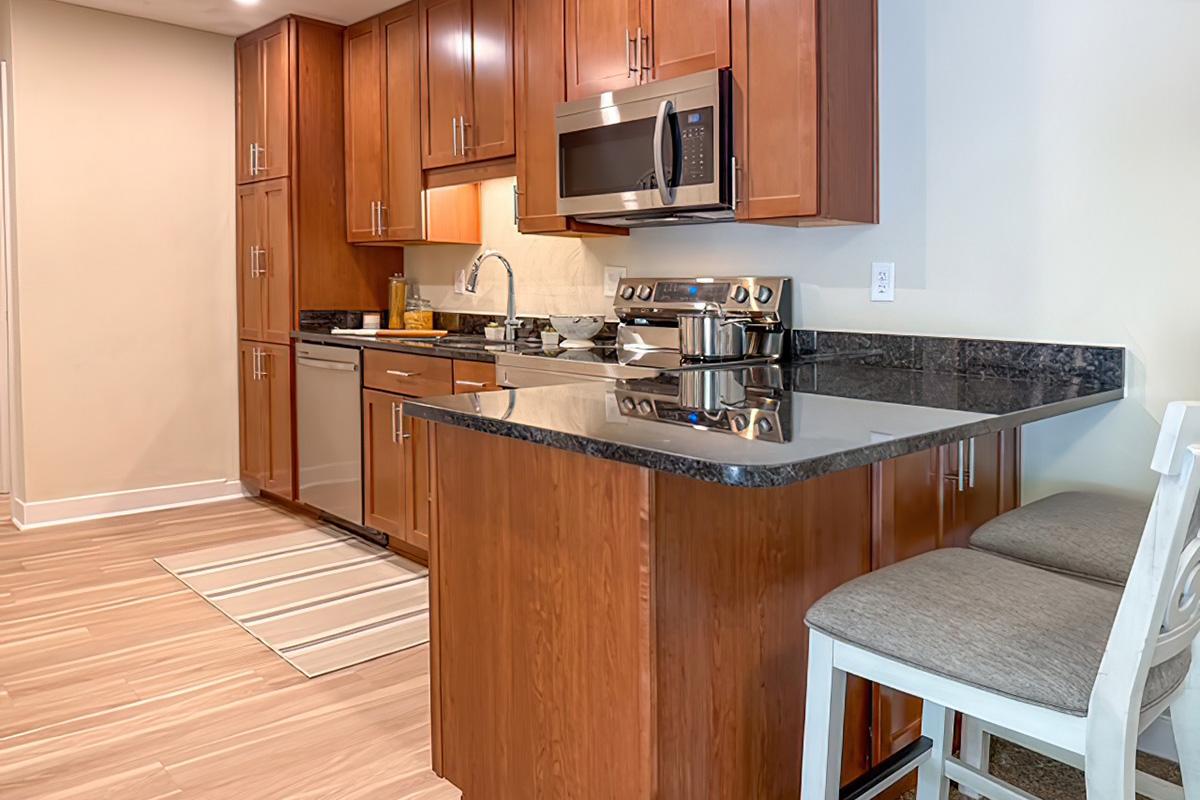 Modern kitchen featuring wooden cabinetry, stainless steel appliances including a microwave and stove, a sleek black countertop, and two bar stools. A light-colored rug is placed on the floor, and the space is well-lit with warm lighting.