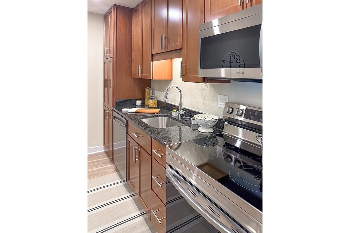 A modern kitchen featuring wooden cabinetry, a stainless steel stove, and a microwave. The countertop is dark granite, with a sink and kitchen accessories. There's a small bowl and a cutting board on the counter, reflecting a clean and organized cooking space. A rug adds warmth to the floor.