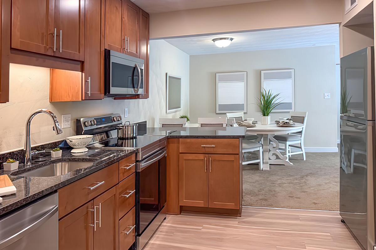 Modern kitchen featuring wood cabinets, stainless steel appliances, and a granite countertop. An open layout connects the kitchen to a dining area with a round table and white chairs, complemented by decorative plants and framed wall art. Soft lighting enhances the inviting atmosphere.