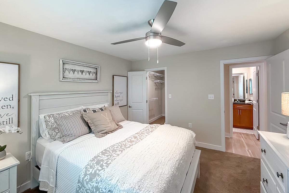 A cozy bedroom featuring a white bed with decorative pillows, a ceiling fan, and light-colored walls. There is a wooden dresser and a framed wall art piece. A doorway leads to a bathroom area in the background, with a closet to the left. The overall atmosphere is calm and inviting.