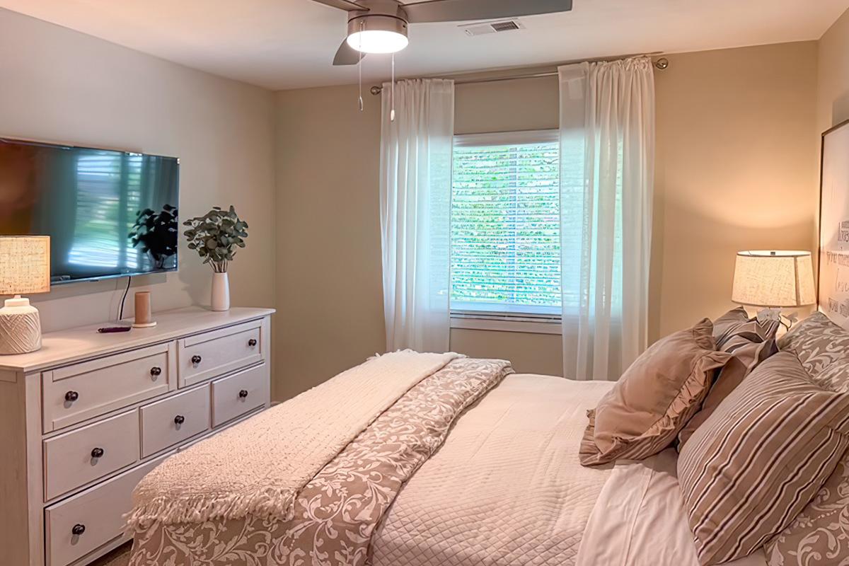 Cozy bedroom with a white bed adorned with light gray and beige bedding. A dresser sits across from the bed, featuring a flat screen TV mounted on the wall. Two bedside lamps illuminate the space, and sheer curtains allow natural light to filter in through the window, creating a warm and inviting atmosphere.