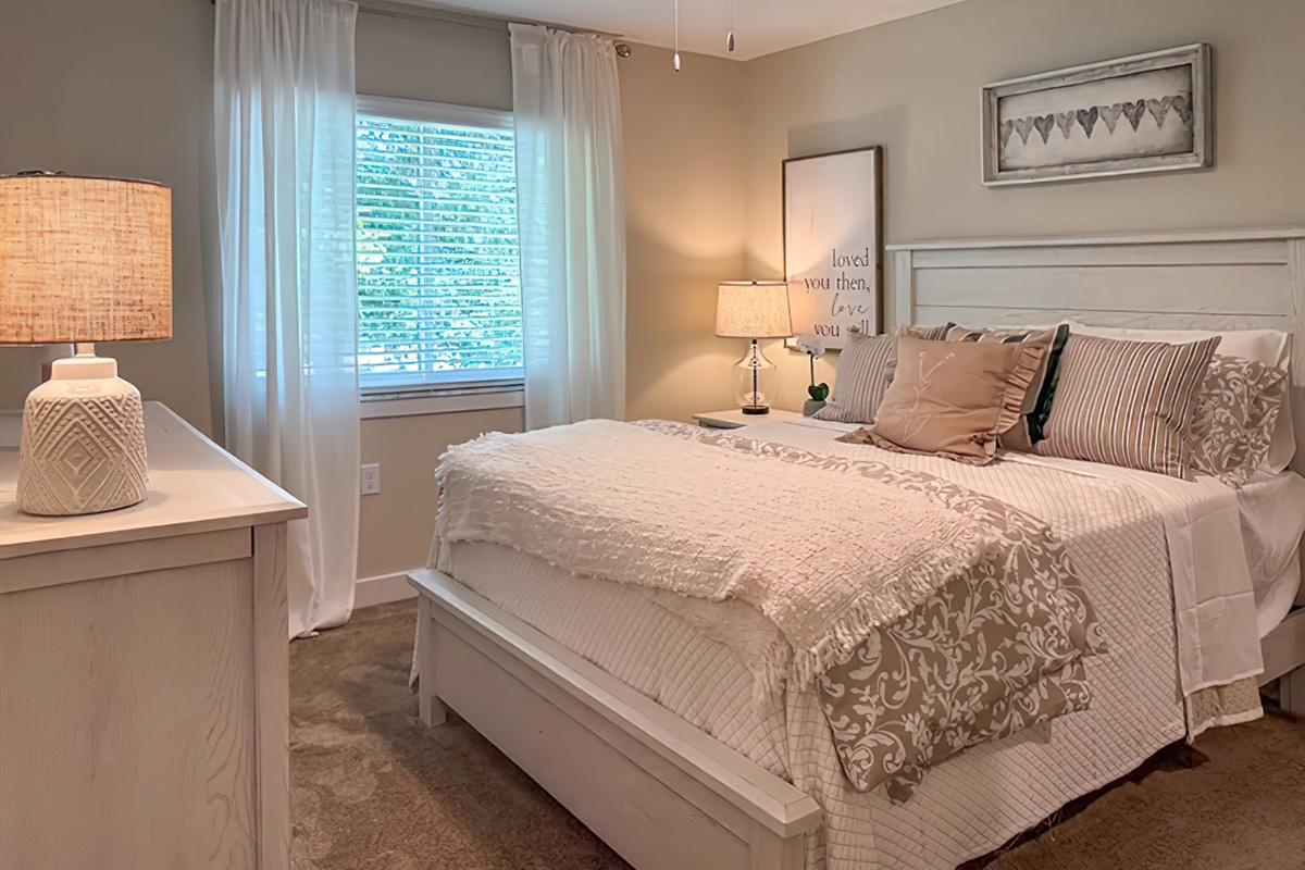 Cozy bedroom featuring a white wooden bed with decorative pillows and a textured throw blanket. A bedside table with a lamp sits next to the bed, and a light-colored dresser is visible. Soft curtains frame a window that lets in natural light, creating a warm and inviting atmosphere.