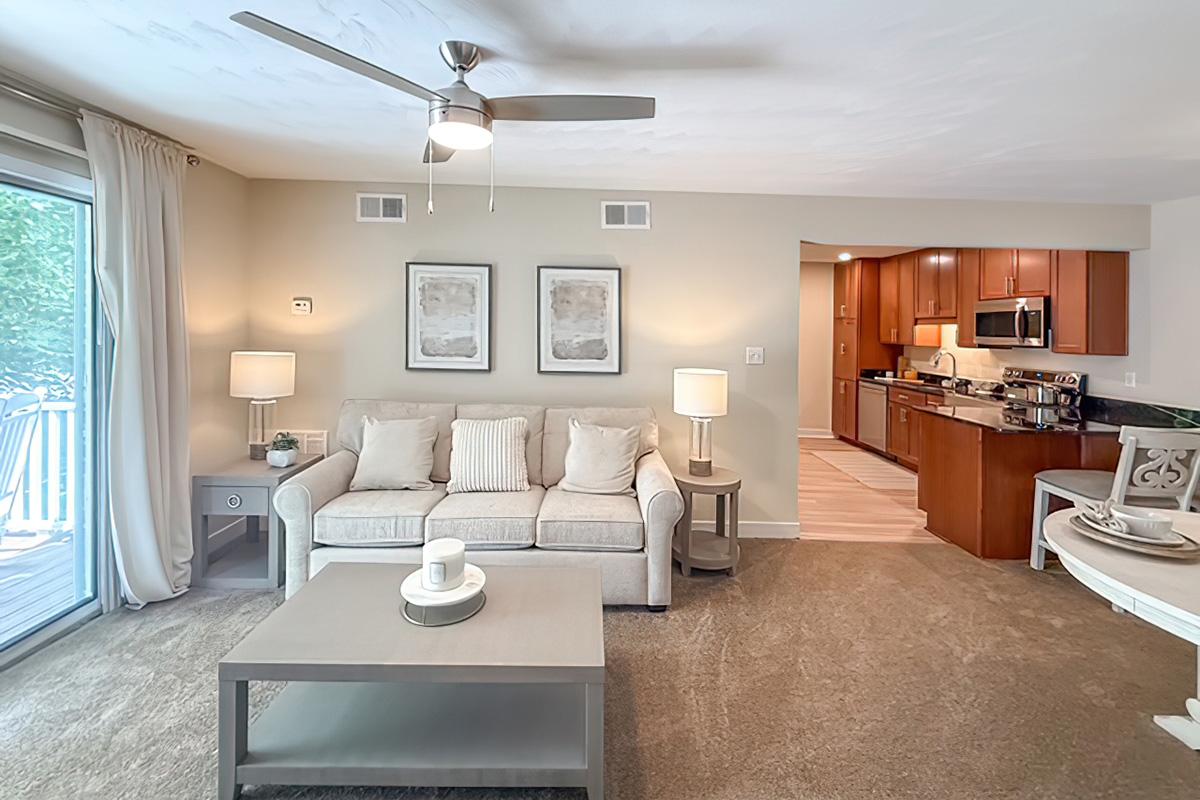 A cozy living room featuring a light-colored sofa with decorative pillows, a coffee table, and two lamps. There are two framed artworks on the wall, and a sliding glass door leads to a patio. The adjacent area shows a modern kitchen with wooden cabinets and stainless steel appliances. Soft lighting and neutral colors create a warm ambiance.