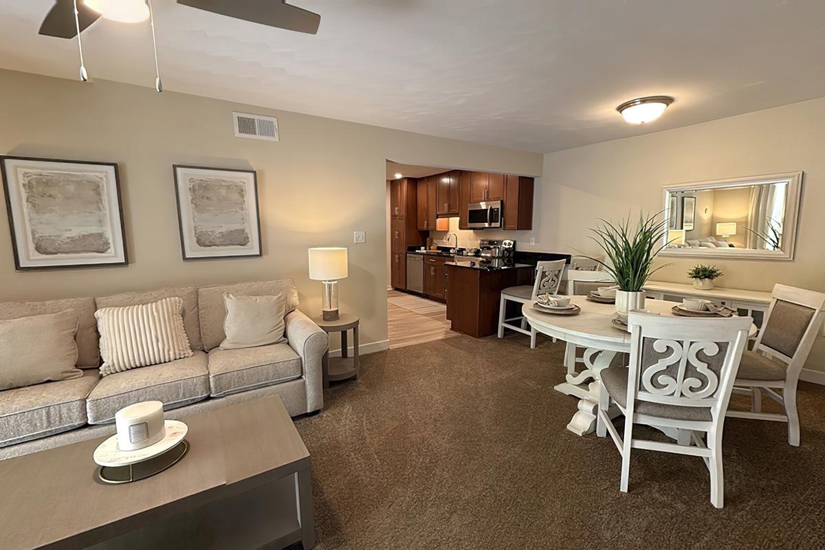 A cozy living room with a light-colored couch, coffee table, and two framed artworks on the wall. A dining area features a white table with four chairs, and a small plant centerpiece. The kitchen is visible in the background, showcasing wooden cabinets and modern appliances. Overall, the space is well-lit and inviting.