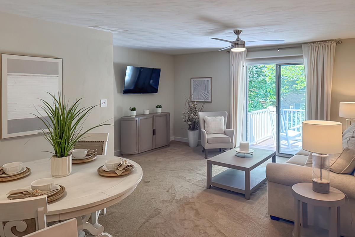 A cozy living room area featuring a light-colored sofa, a small coffee table, and a dining table set for four. There's a flat-screen TV mounted on the wall, and large glass doors leading to a patio with greenery visible outside. The space is well-lit with a ceiling fan and neutral decor.