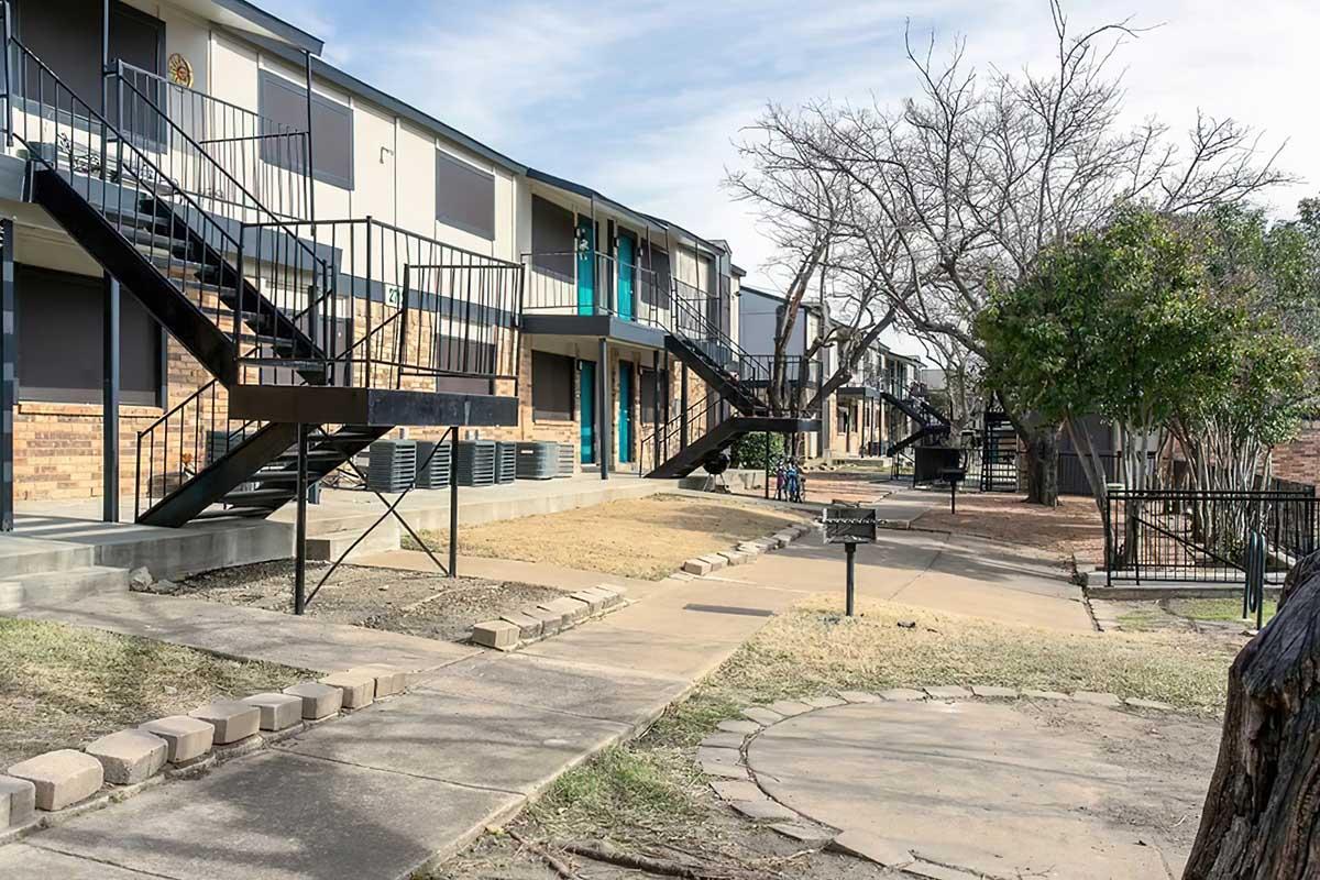View of a residential apartment complex featuring two-story buildings with black metal staircases. The pathway is lined with bare trees and grass. There are picnic areas with grills and air conditioning units. The sky is partly cloudy, creating a calm, suburban atmosphere.