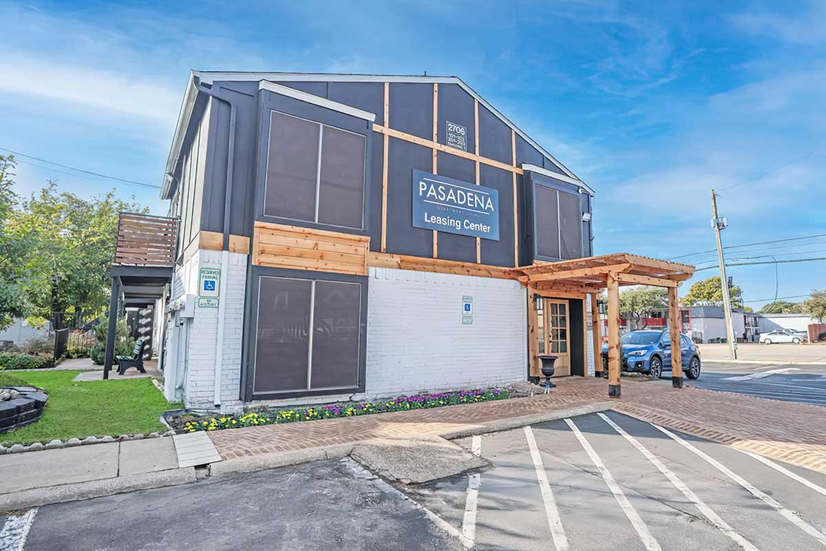 A two-story leasing center building with a modern exterior design featuring a combination of dark panels and white brick. The entrance has a wooden awning and landscaped flower beds. Parking spaces are visible in front, with a blue car parked nearby and a clear blue sky above.