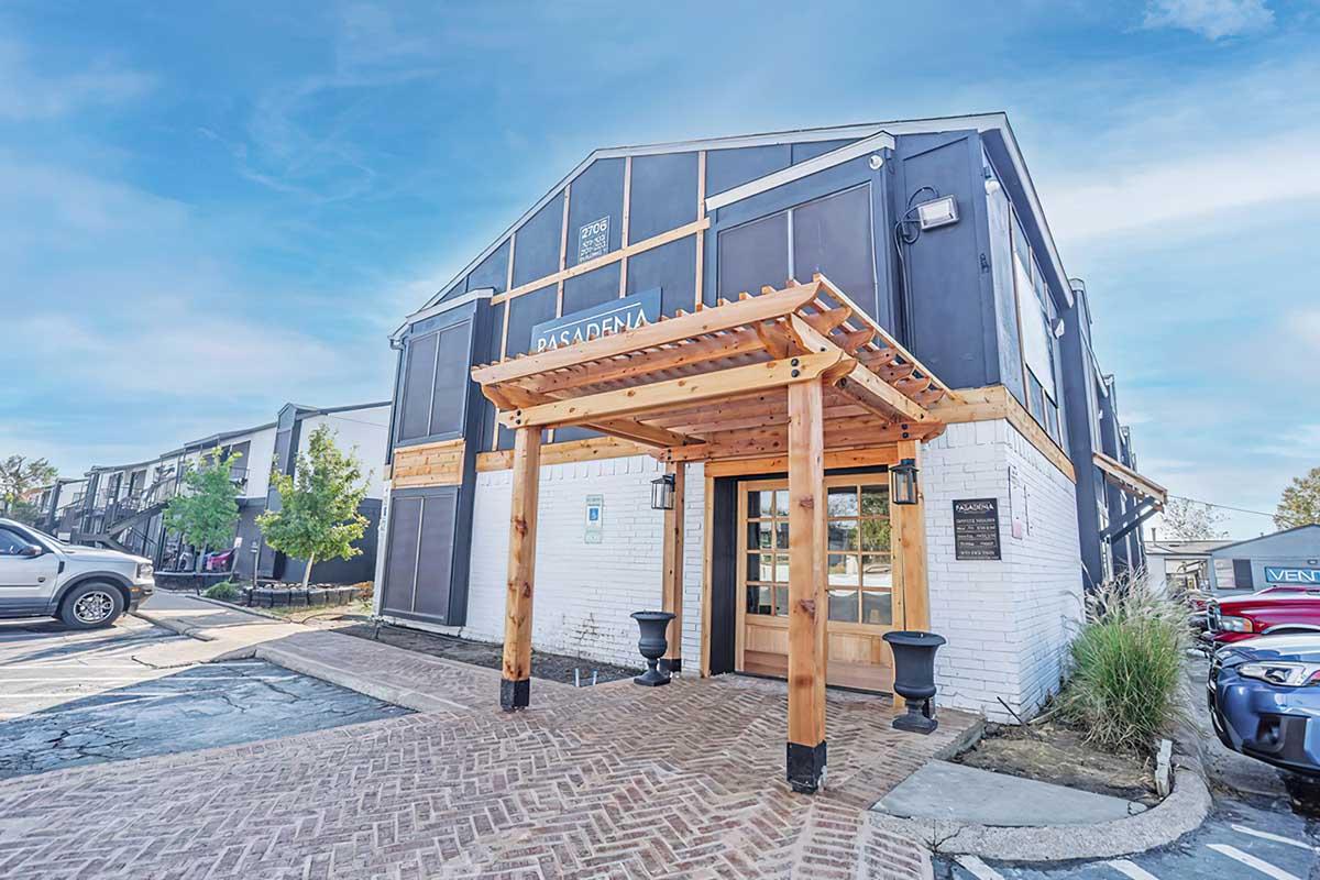 Exterior view of a modern building with a wooden entrance and signage for "Pasadena." The building features a combination of dark and white siding, surrounded by greenery and parked cars. A brick pathway leads to the entrance, showcasing a clean and inviting atmosphere.