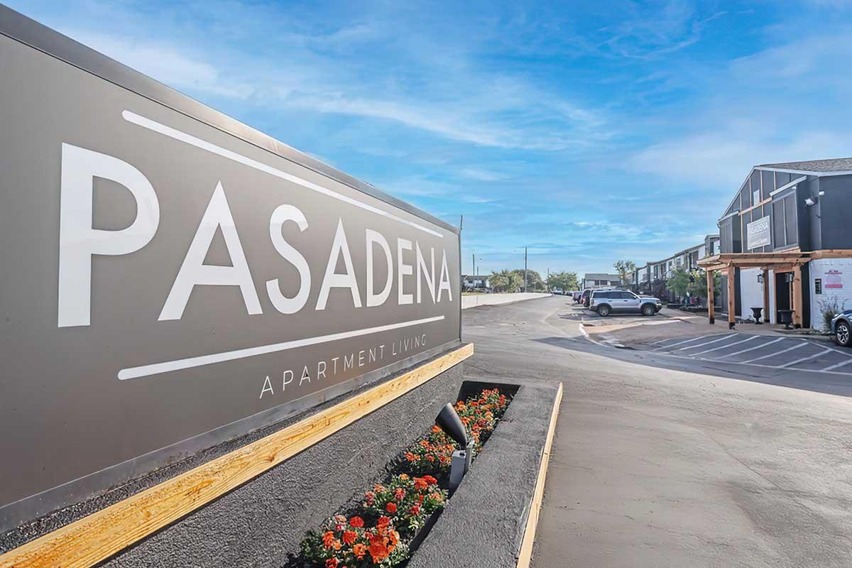 Sign for Pasadena Apartment Living with a landscaped flower bed in front and buildings visible in the background. The sky is clear and blue, creating a welcoming atmosphere for potential residents.