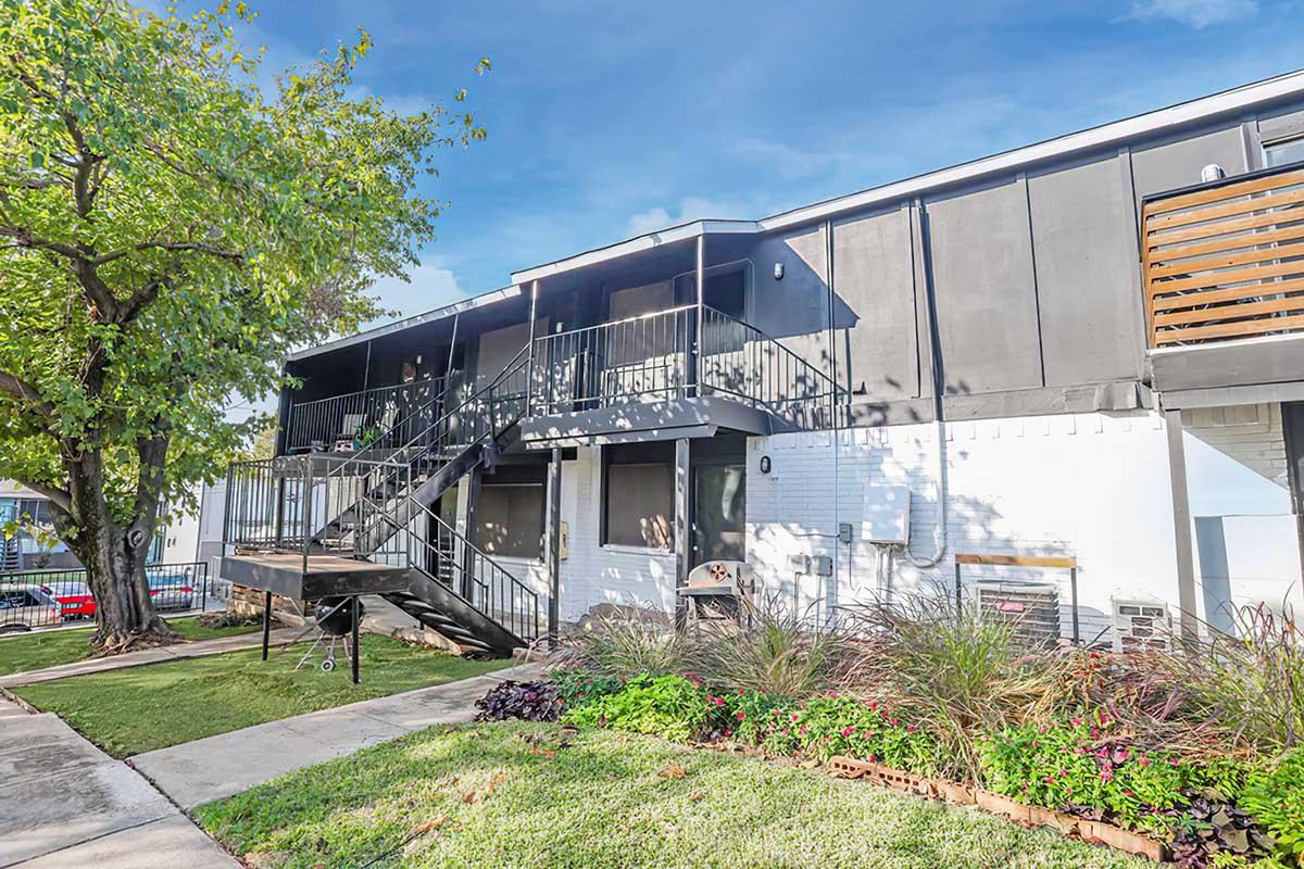 A two-story apartment building with a black exterior and a staircase leading to the upper level. There is a lush green lawn in front, featuring flowerbeds and shrubs. A tree provides shade to the area, and there are outdoor air conditioning units visible. The sky is clear and bright.