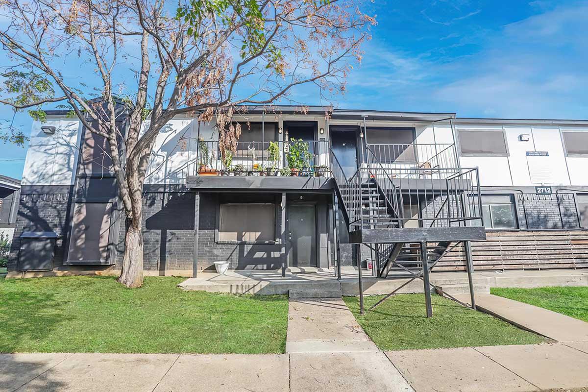 Two-story apartment building with a tree in front. The exterior features a combination of dark and light paint, with a staircase leading to the upper level. The ground level has a door and a small porch area. The surrounding lawn is well-maintained with a clear blue sky above.
