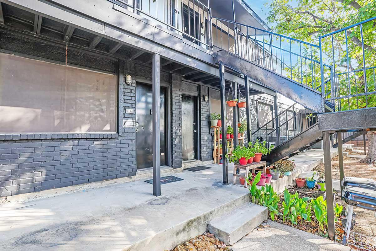 A view of a multi-story building with a black metal staircase on the side. The entrance features a porch with potted plants and greenery, surrounded by a concrete pathway. Windows above the entrance are covered, and there is a sense of inviting outdoor space.